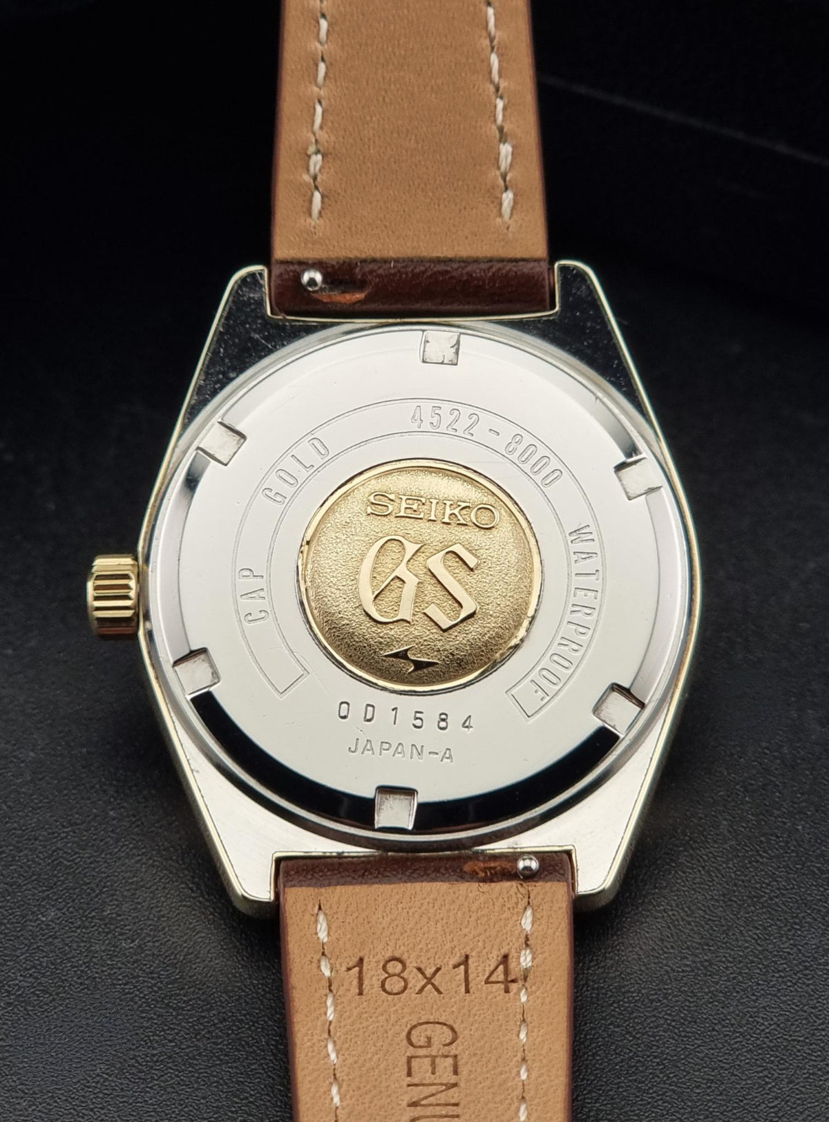 Close-up of the back of a Vintage Grand Seiko watch 4522-8000 with a silver-toned case, gold-tone center detail, and a brown leather strap on a dark background.