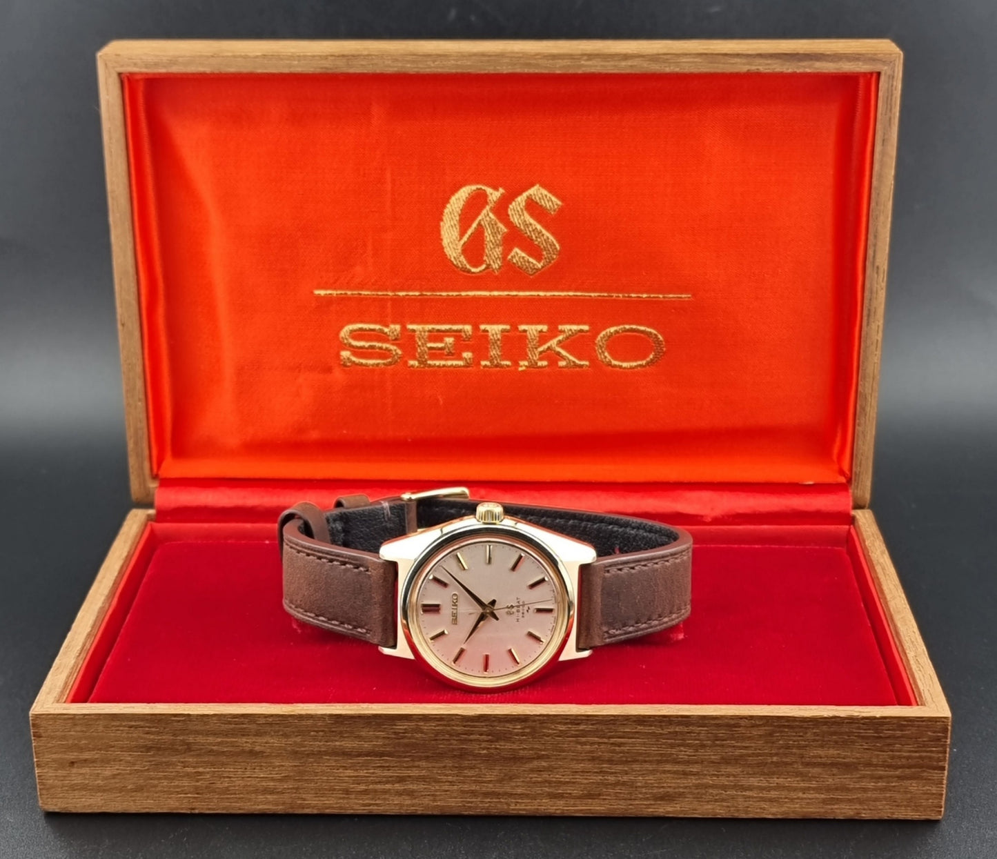 Vintage Grand Seiko Gold 4520-8000 wristwatch with a brown leather strap on a dark textured background in its box