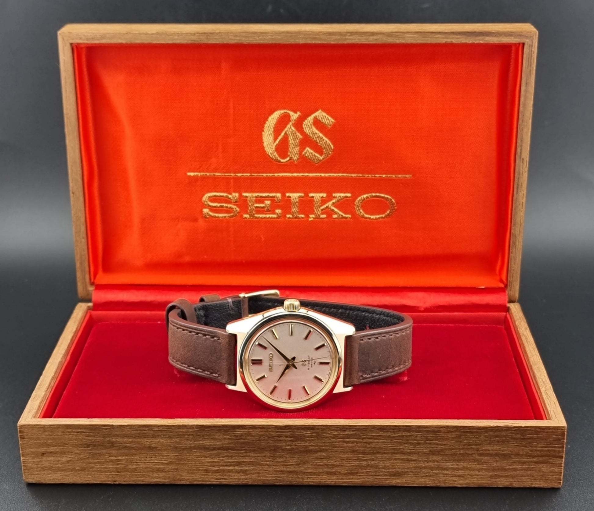 Vintage Grand Seiko Gold 4520-8000 wristwatch with a brown leather strap on a dark textured background in its box