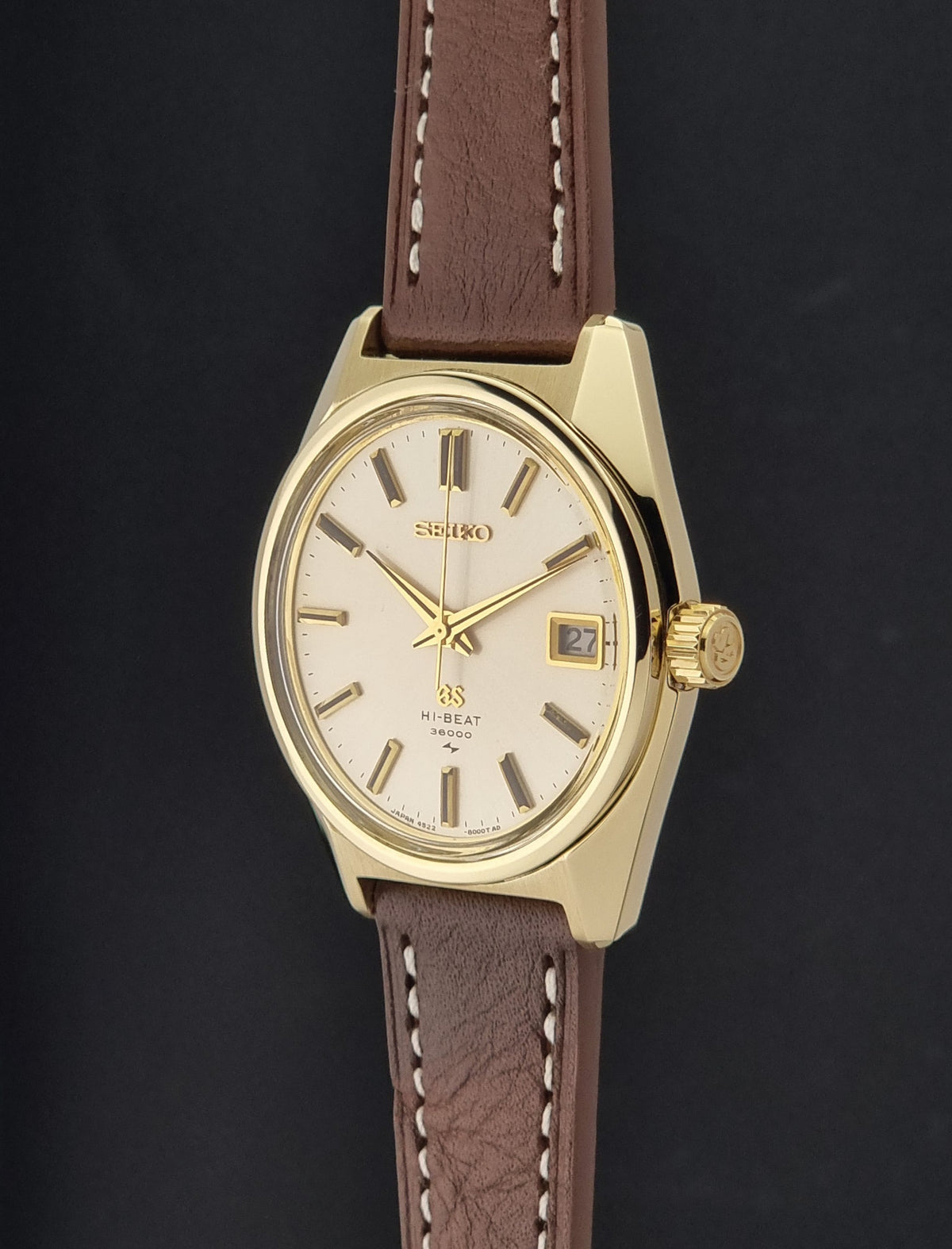 Vintage Grand Seiko watch 4522-8000 with a gold-tone case, cream dial, and brown leather strap with white stitching, shown against a dark background.