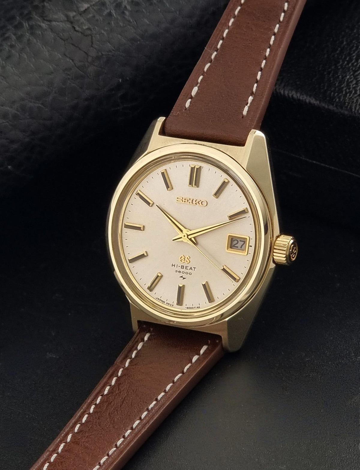 Vintage Grand Seiko watch 4522-8000 with a gold-tone case, cream dial, and brown leather strap with white stitching, displayed diagonally on a dark textured surface.