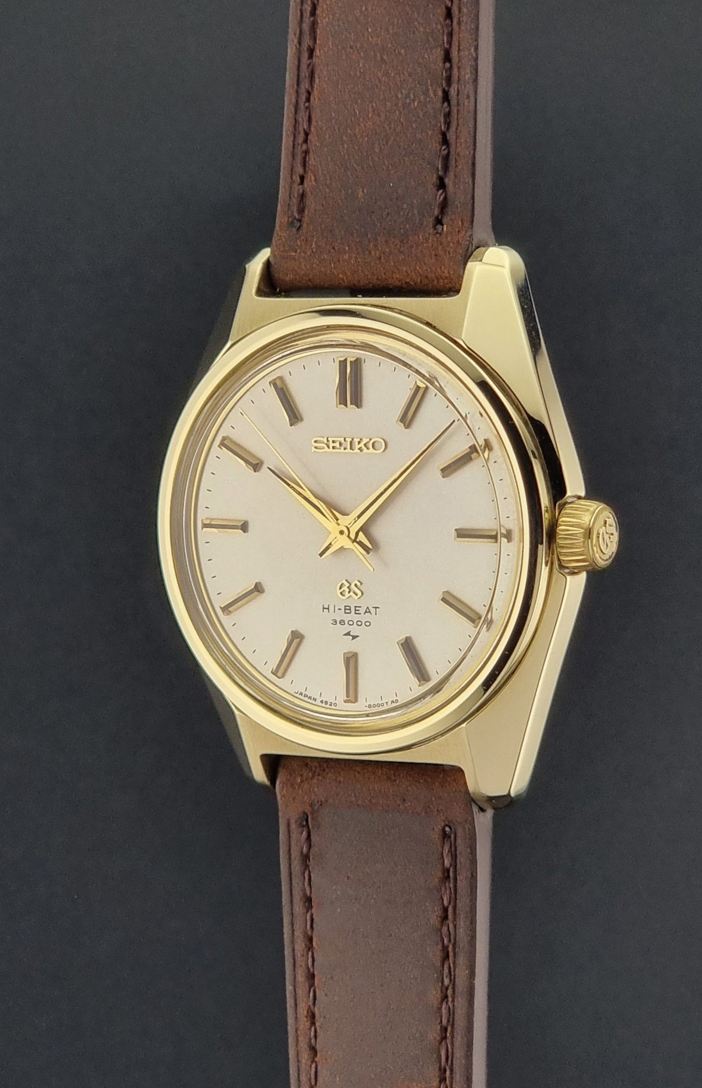 Vintage Grand Seiko Gold 4520-8000 wristwatch with a brown leather strap on a dark textured background crown side profile