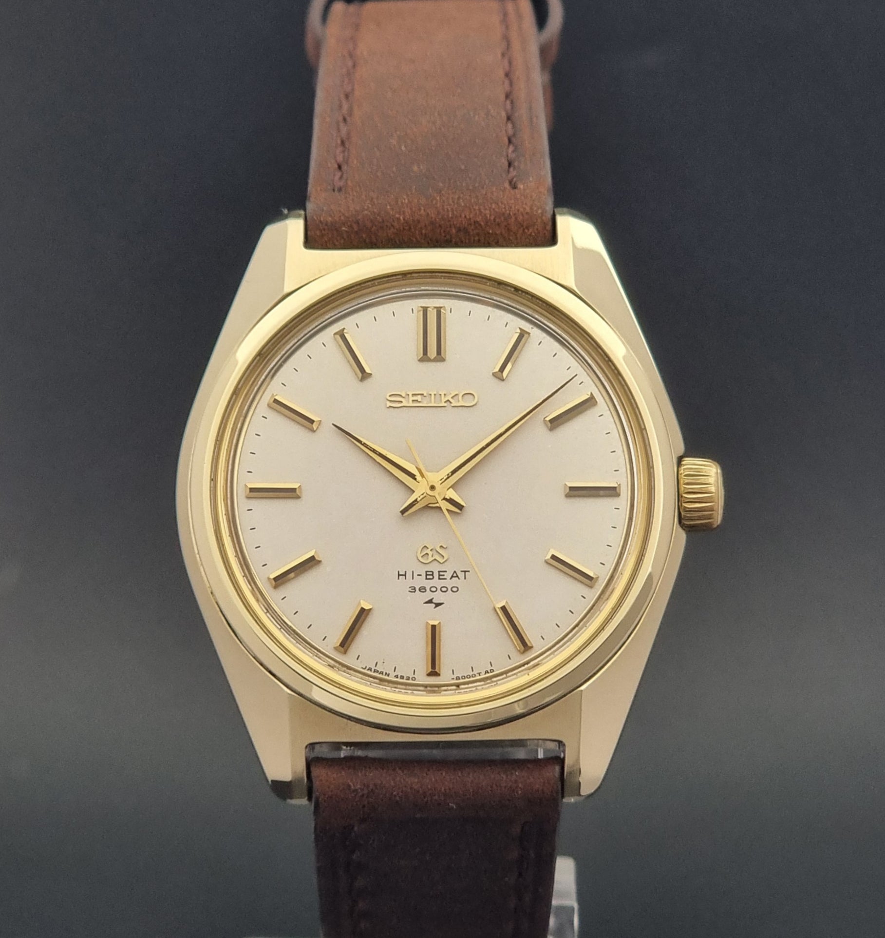 Vintage Grand Seiko Gold 4520-8000 wristwatch with a brown leather strap on a dark textured background front view