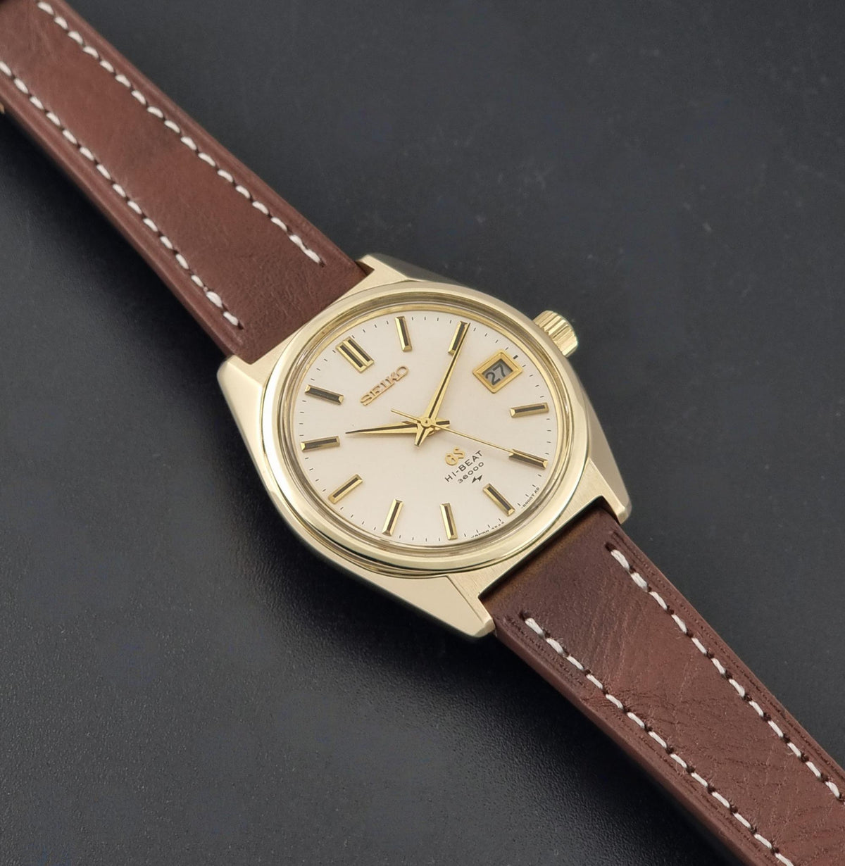 Vintage Grand Seiko 4522-8000 wristwatch with a gold-toned case, cream-colored dial, and a brown leather strap with white stitching, shown diagonally on a dark textured background.