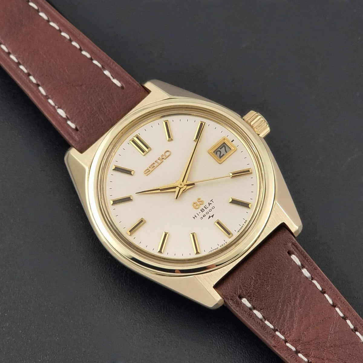 Vintage Grand Seiko watch 4522-8000 with a gold-tone case, cream dial, and brown leather strap, shown diagonally on a dark textured background.