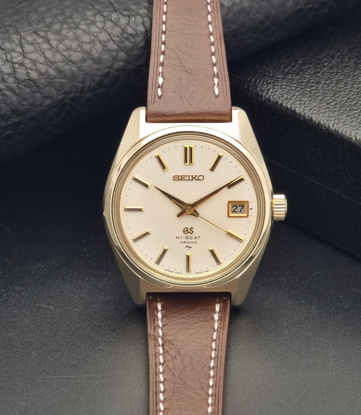 Vintage Grand Seiko watch 4522-8000 with a gold-tone case, cream dial, and brown leather strap, displayed on a textured black surface.