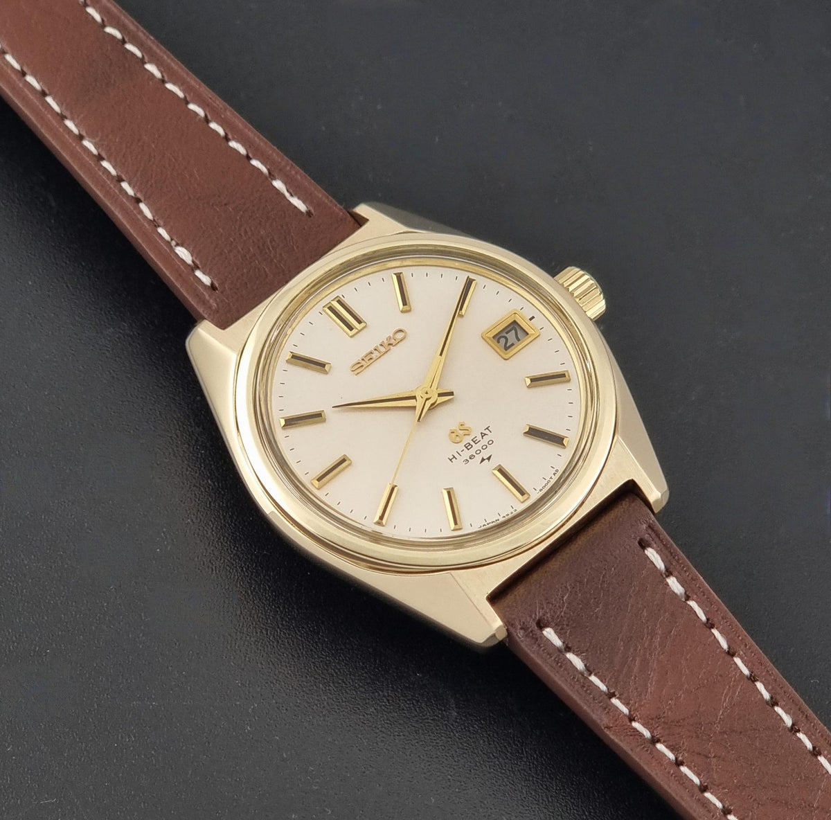 Vintage Grand Seiko watch 4522-8000 with a gold-tone case, cream-colored dial, and a brown leather strap with white stitching, shown diagonally on a dark textured background.