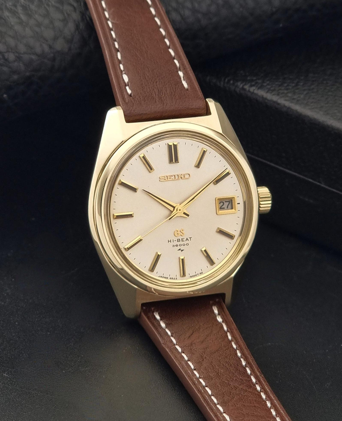 Vintage Grand Seiko watch 4522-8000 with a gold-toned case, cream dial, and brown leather strap with white stitching, photographed on a dark textured surface.