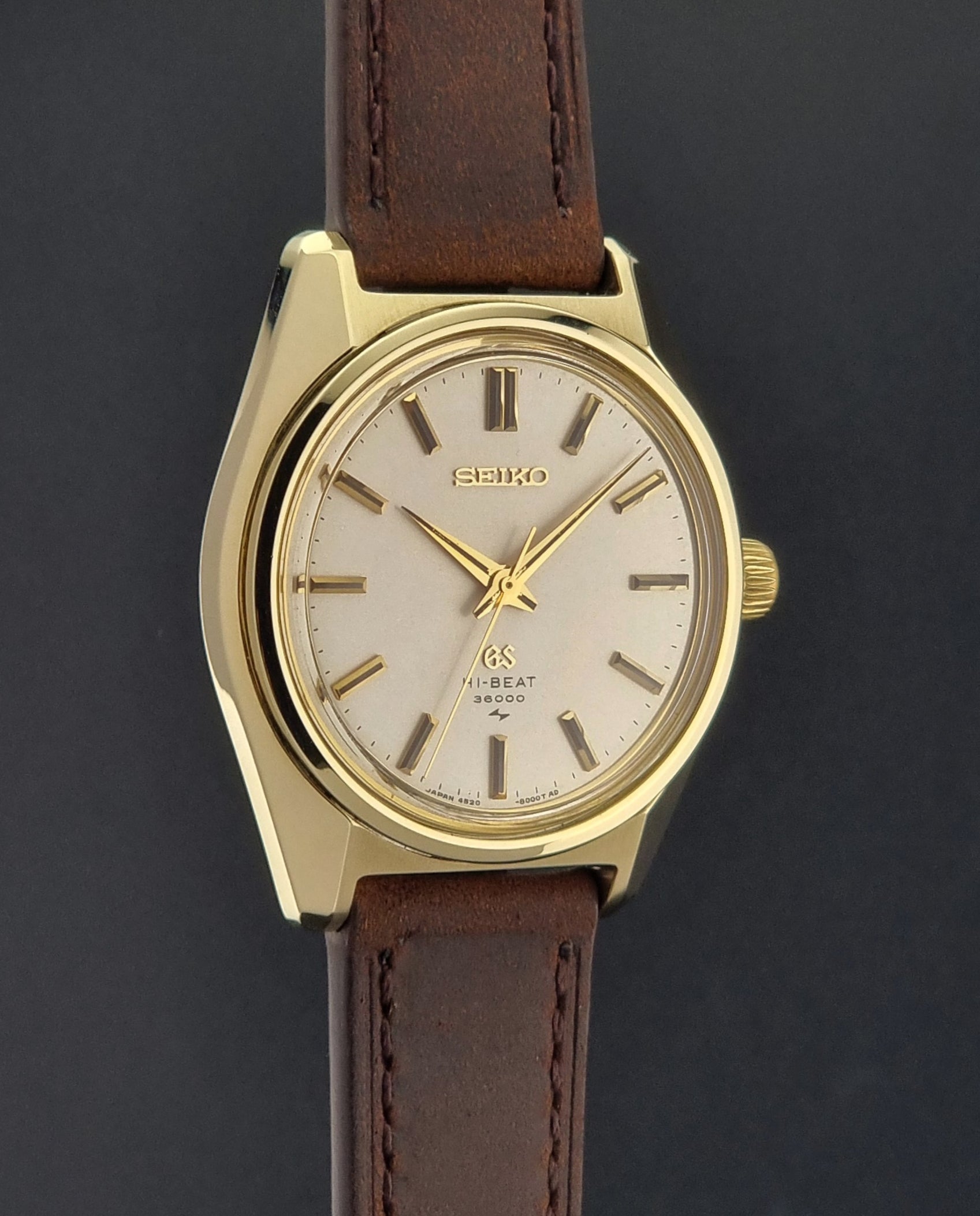 Vintage Grand Seiko Gold 4520-8000 wristwatch with a brown leather strap on a dark textured background side profile