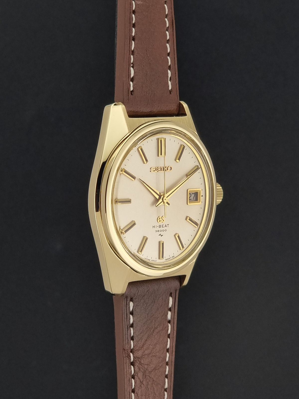 Vintage Grand Seiko watch 4522-8000 with a gold-tone case, cream-colored dial, and a brown leather strap with white stitching, shown against a dark background.