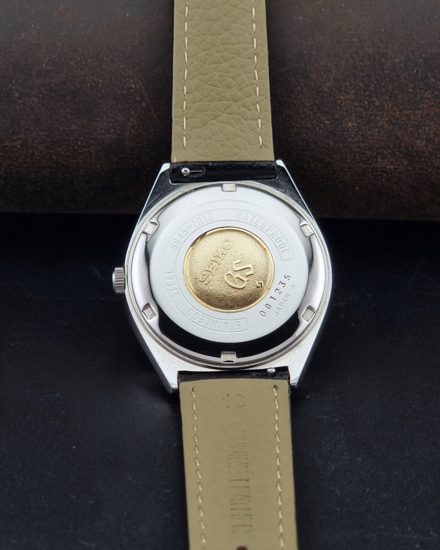 Vintage Grand Seiko Wristwatch 5645-7010 with a silver-toned round case, white dial, and black textured leather strap, on a dark gray background case back view