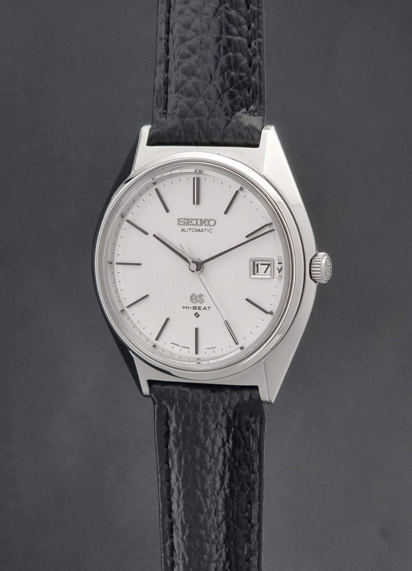 Vintage Grand Seiko Wristwatch 5645-7010 with a silver-toned round case, white dial, and black textured leather strap, on a dark gray background crown profile view