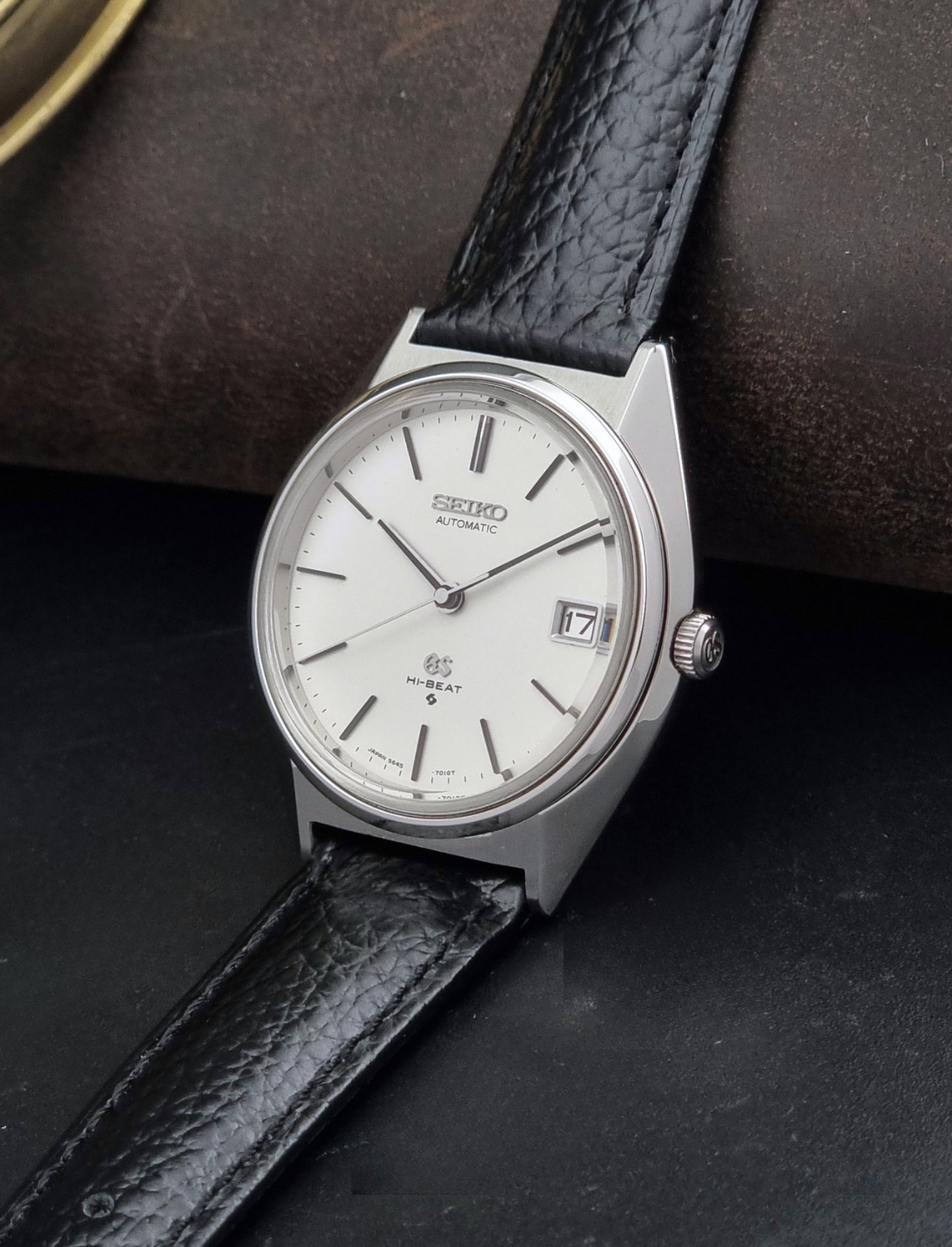 Vintage Grand Seiko Wristwatch 5645-7010 with a silver-toned round case, white dial, and black textured leather strap, on a dark gray background crown view