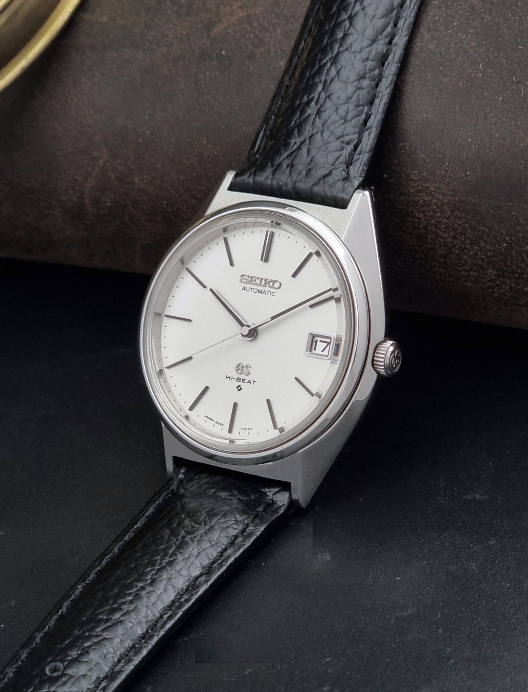 Vintage Grand Seiko Wristwatch 5645-7010 with a silver-toned round case, white dial, and black textured leather strap, on a dark gray background crown view