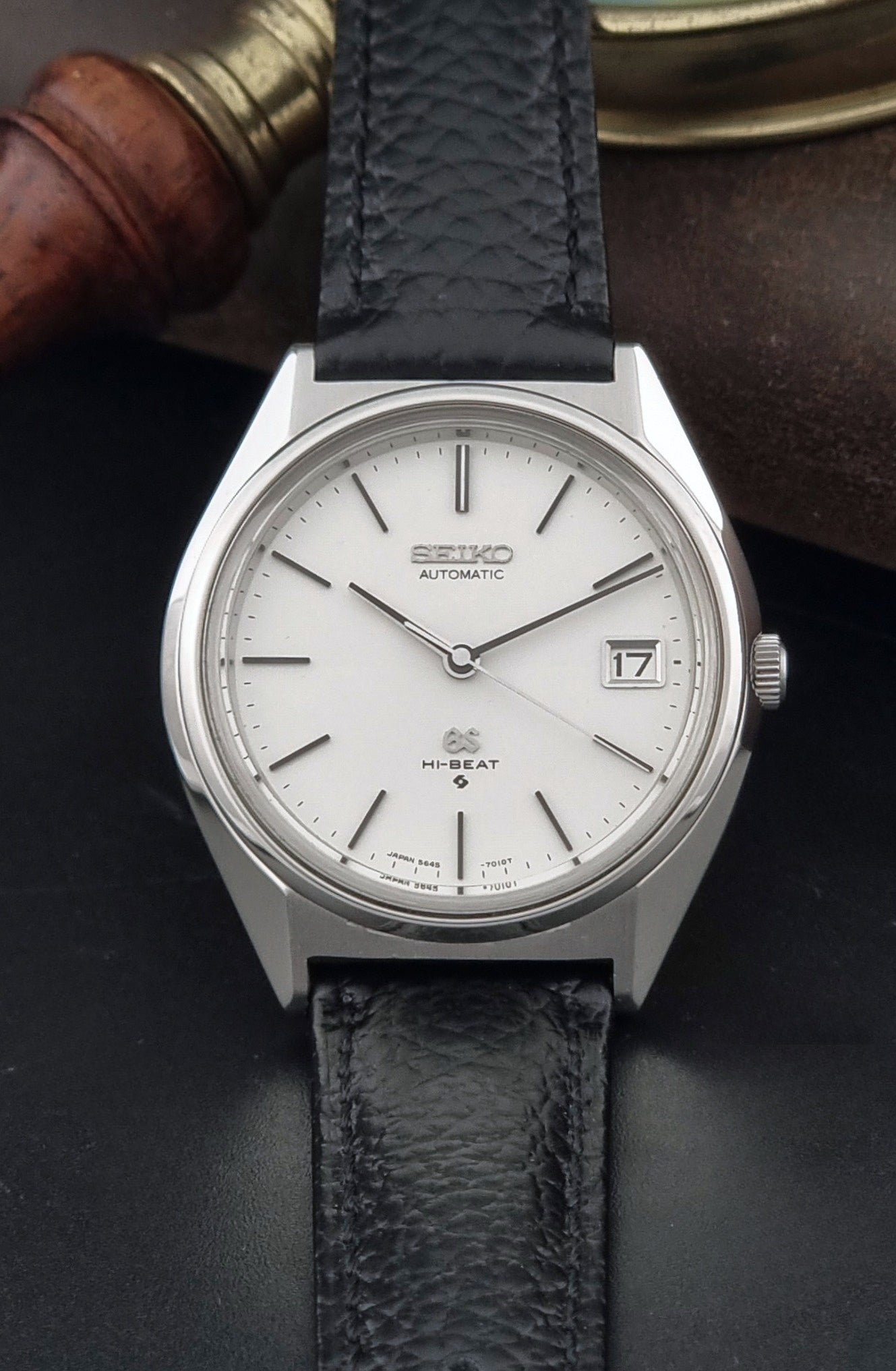 Vintage Grand Seiko Wristwatch 5645-7010 with a silver-toned round case, white dial, and black textured leather strap, on a dark gray background close dial view