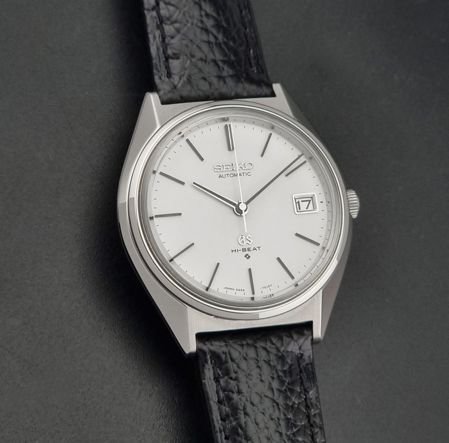 Vintage Grand Seiko Wristwatch 5645-7010 with a silver-toned round case, white dial, and black textured leather strap, on a dark gray background front view