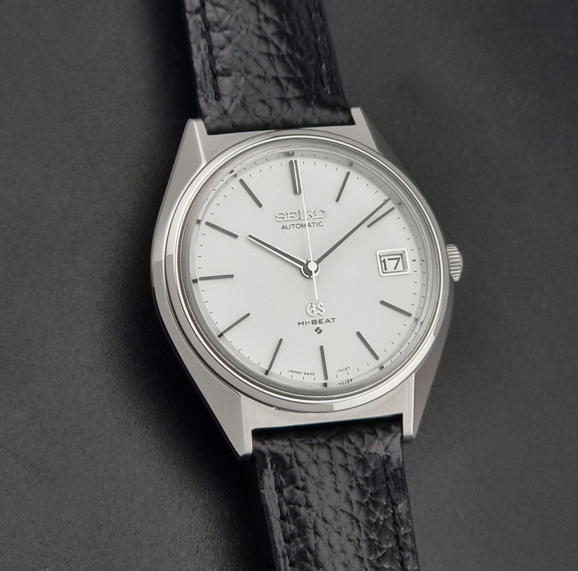 Vintage Grand Seiko Wristwatch 5645-7010 with a silver-toned round case, white dial, and black textured leather strap, on a dark gray background front view