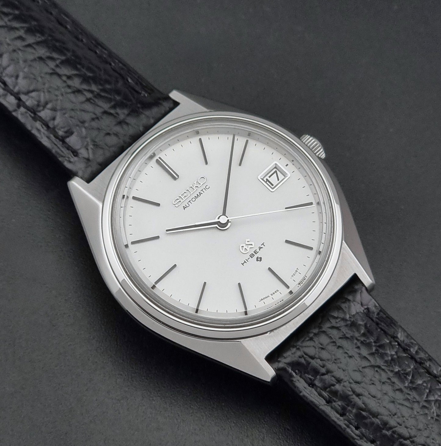 Vintage Grand Seiko Wristwatch 5645-7010 with a silver-toned round case, white dial, and black textured leather strap, on a dark gray background close up dial view