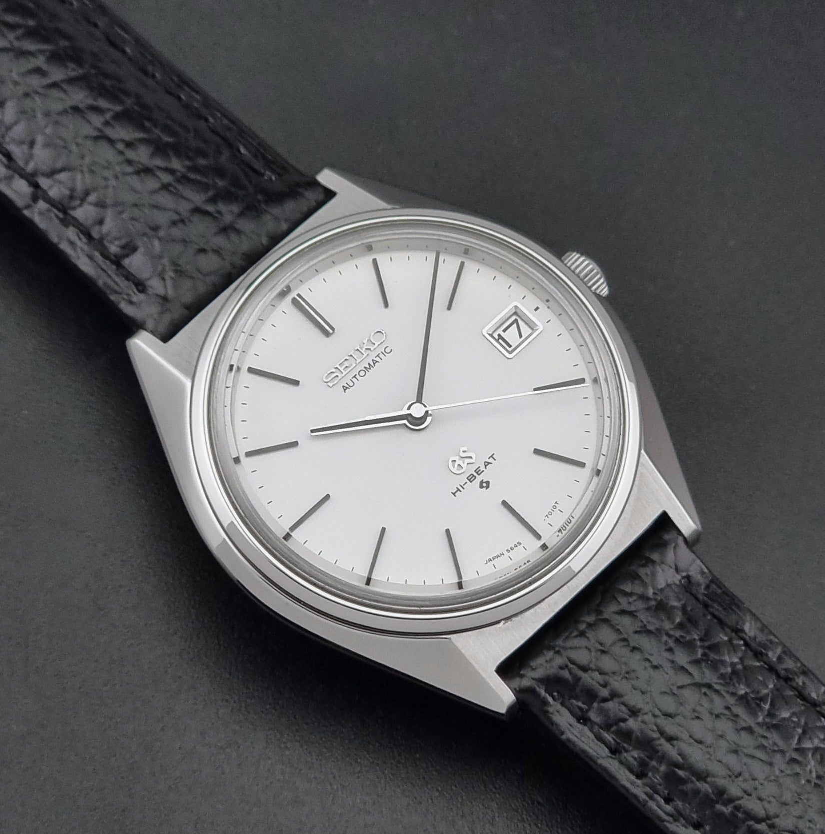 Vintage Grand Seiko Wristwatch 5645-7010 with a silver-toned round case, white dial, and black textured leather strap, on a dark gray background close up dial view