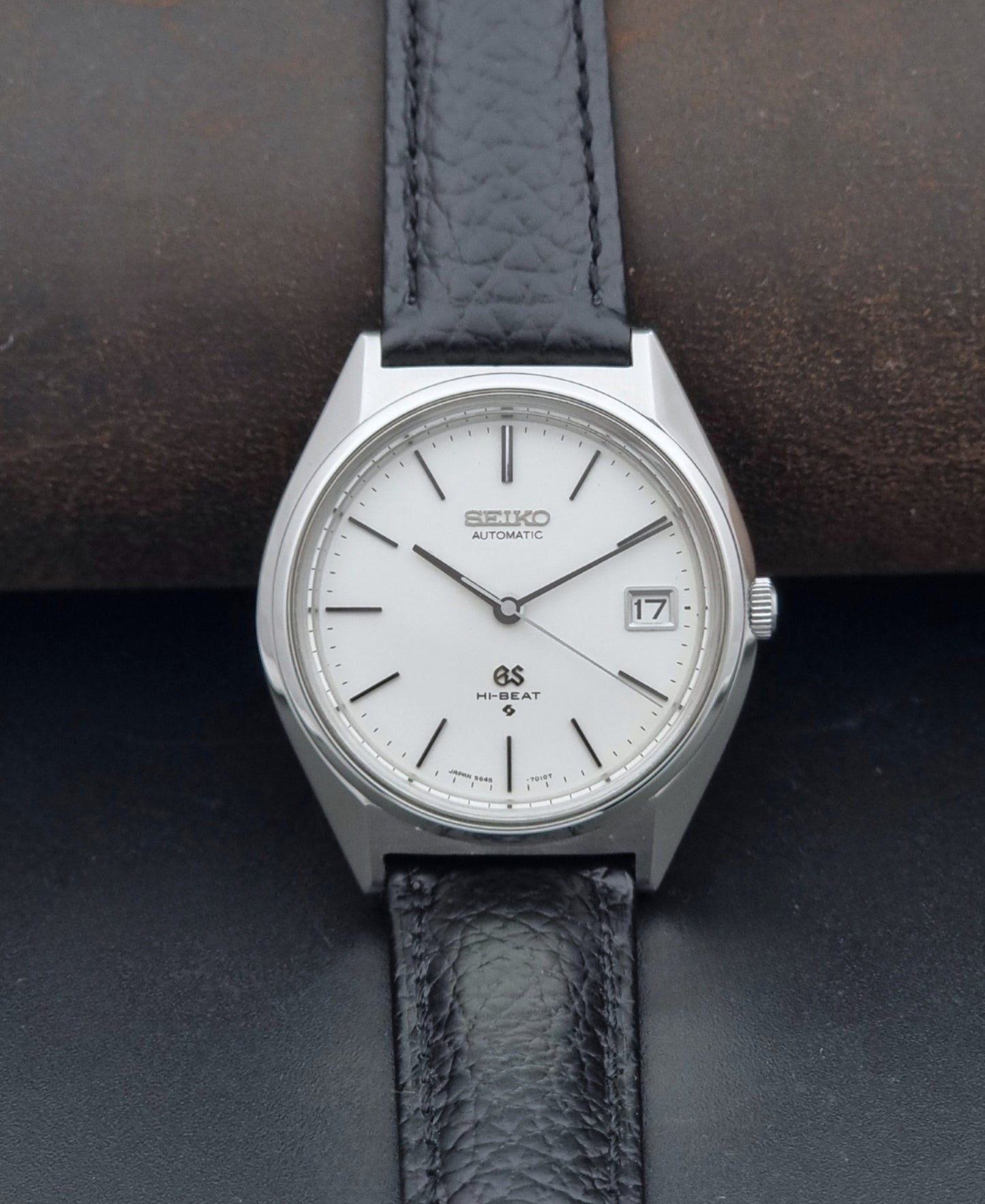 Vintage Grand Seiko Wristwatch 5645-7010 with a silver-toned round case, white dial, and black textured leather strap, on a dark gray background dial view