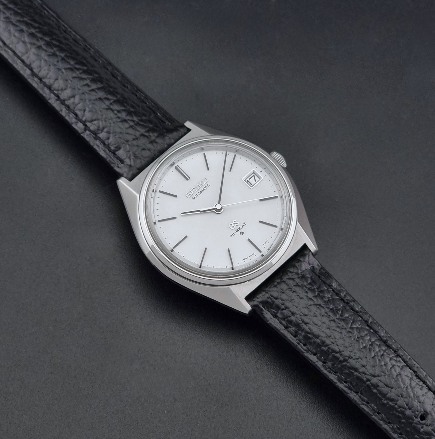 Vintage Grand Seiko Wristwatch 5645-7010 with a silver-toned round case, white dial, and black textured leather strap, on a dark gray background dial
