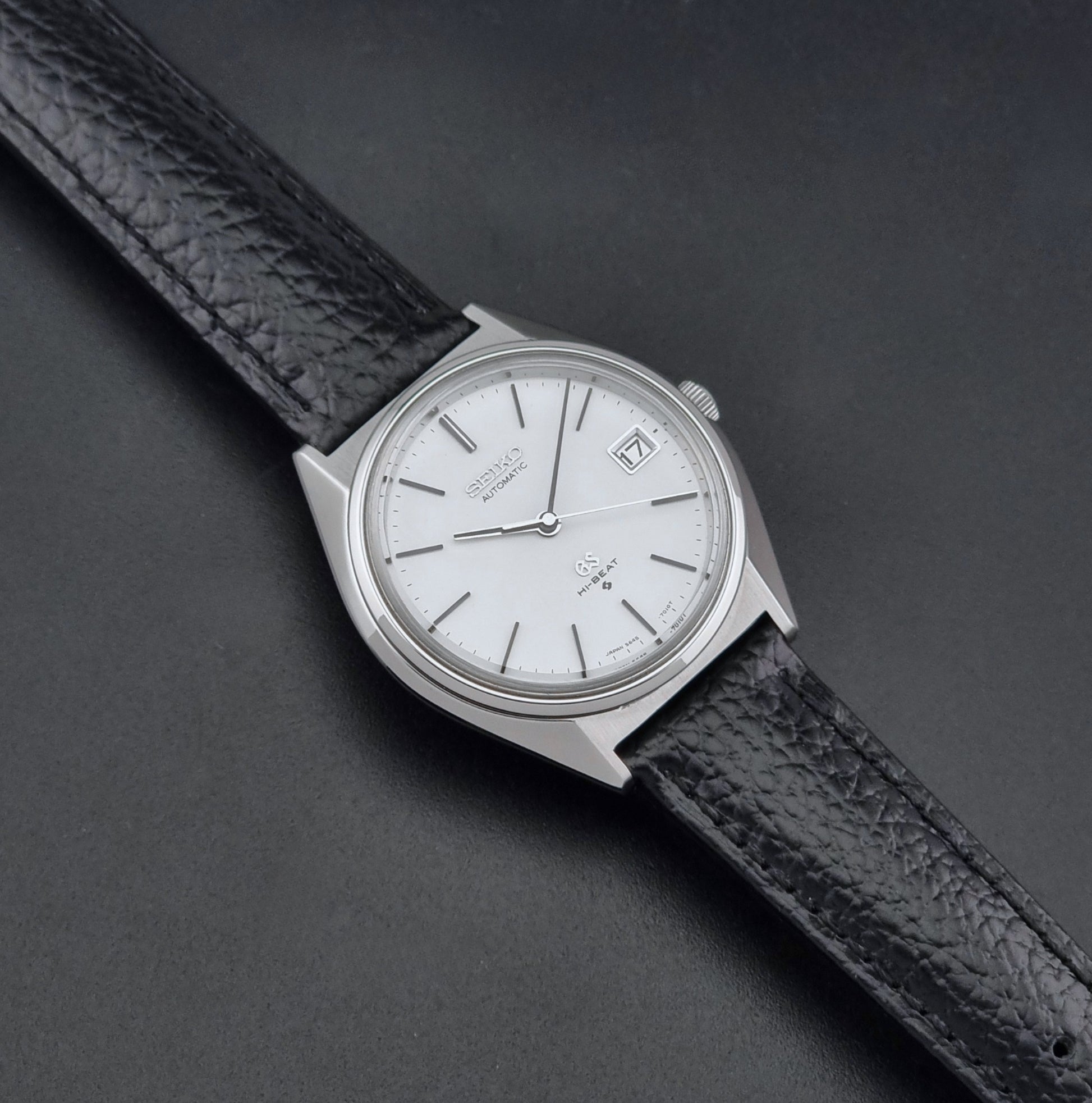 Vintage Grand Seiko Wristwatch 5645-7010 with a silver-toned round case, white dial, and black textured leather strap, on a dark gray background dial