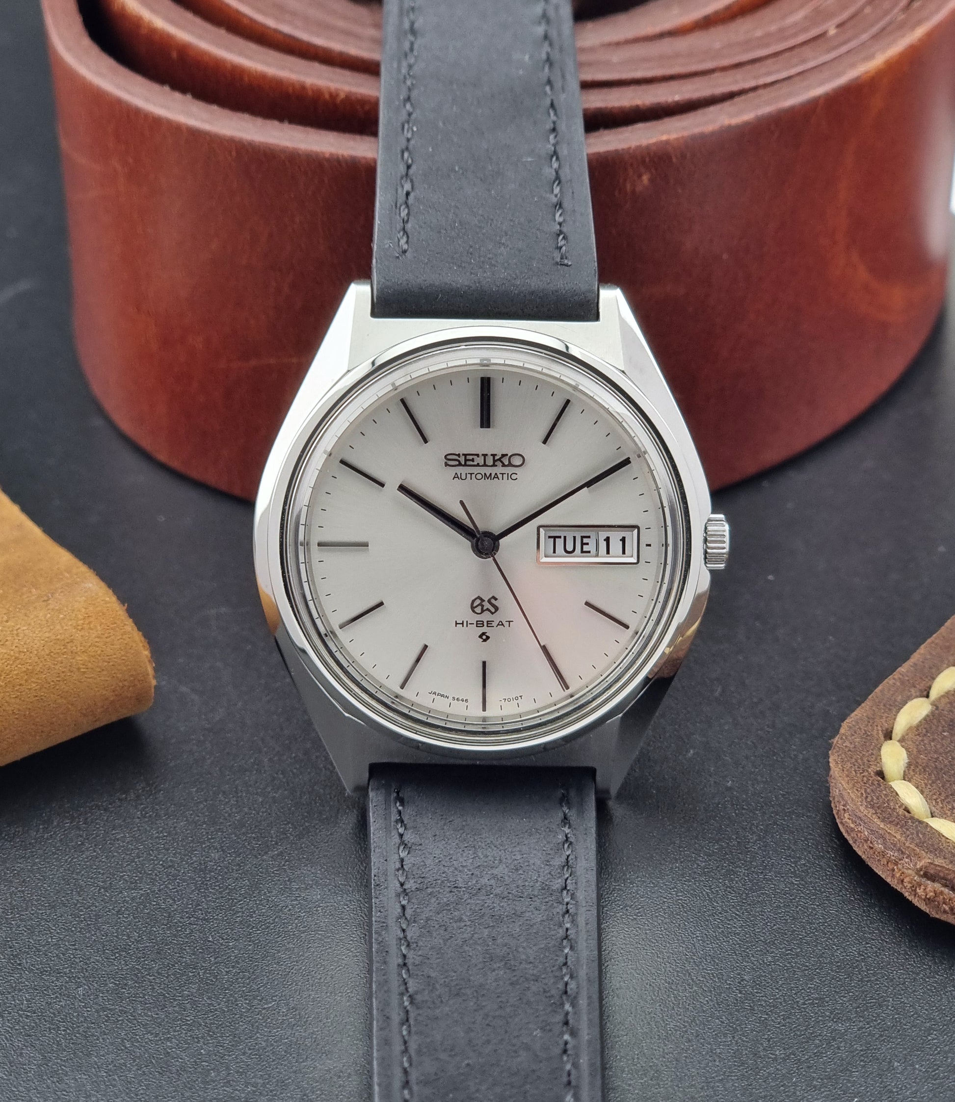 Vintage Grand Seiko watch with black strap on a dark surface with leather items in the background