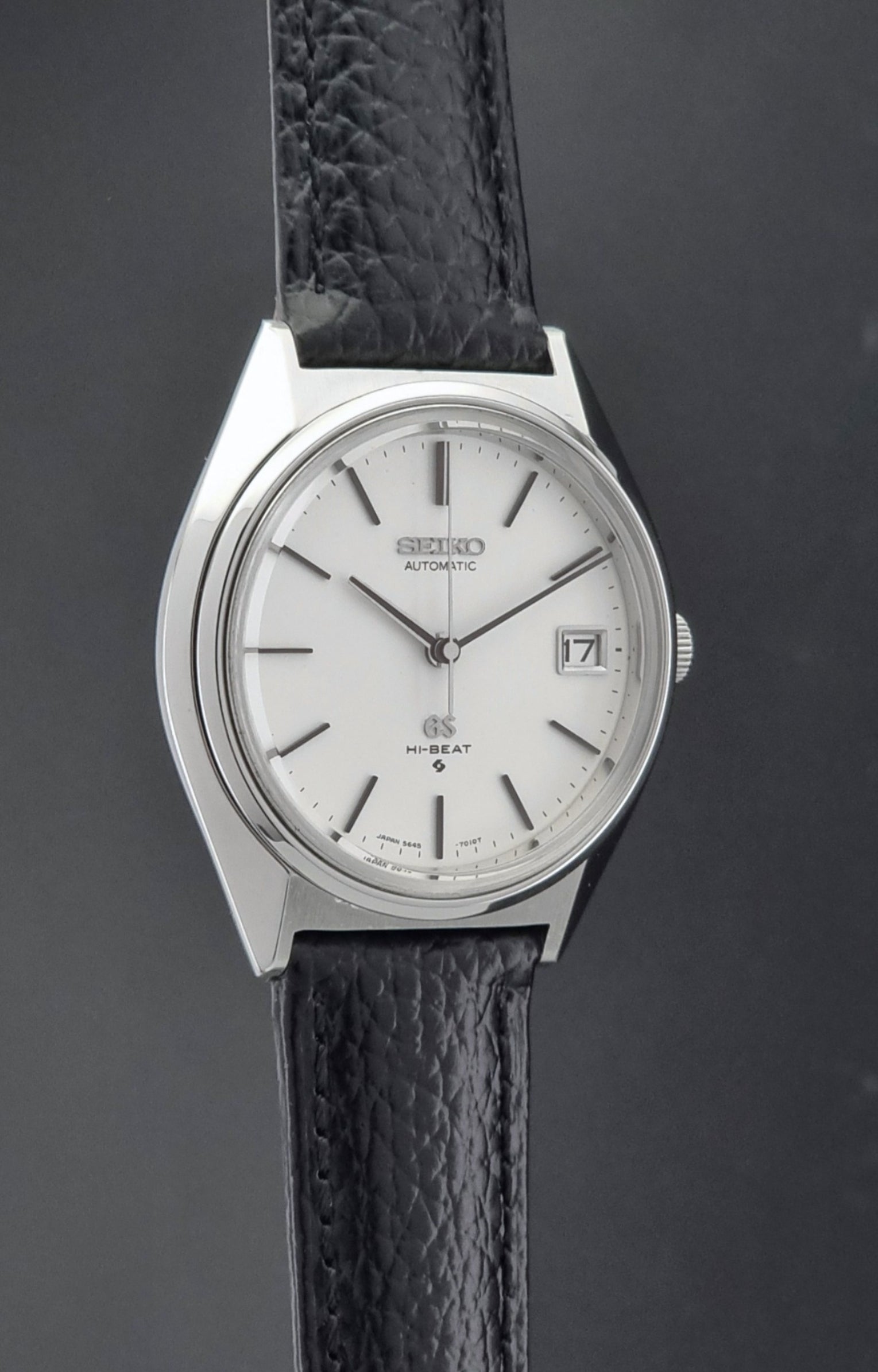 Vintage Grand Seiko Wristwatch 5645-7010 with a silver-toned round case, white dial, and black textured leather strap, on a dark gray background side profile view