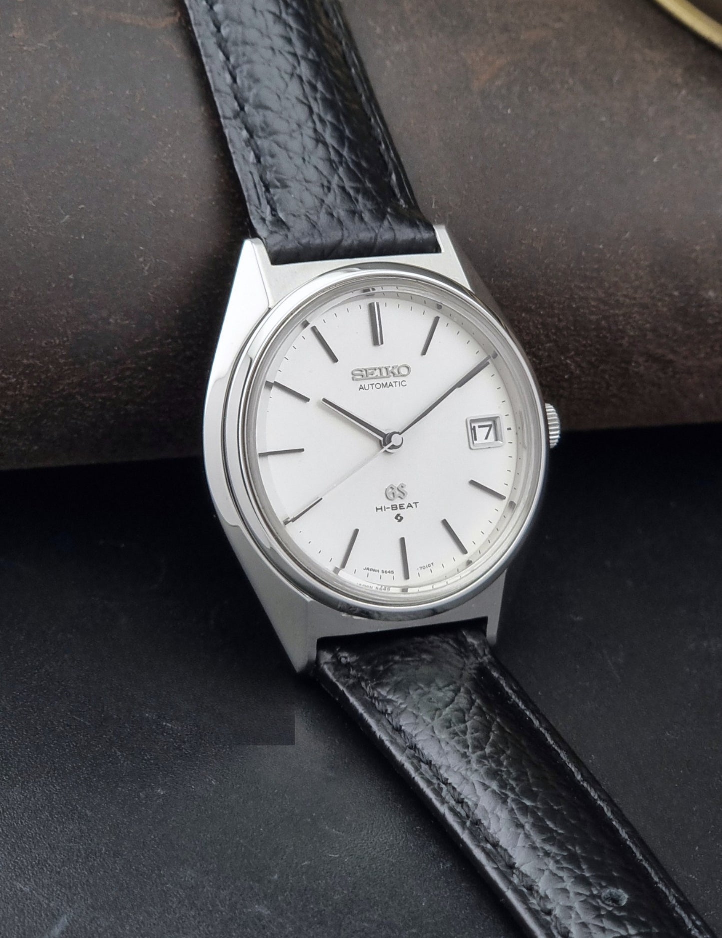 Vintage Grand Seiko Wristwatch 5645-7010 with a silver-toned round case, white dial, and black textured leather strap, on a dark gray background side view