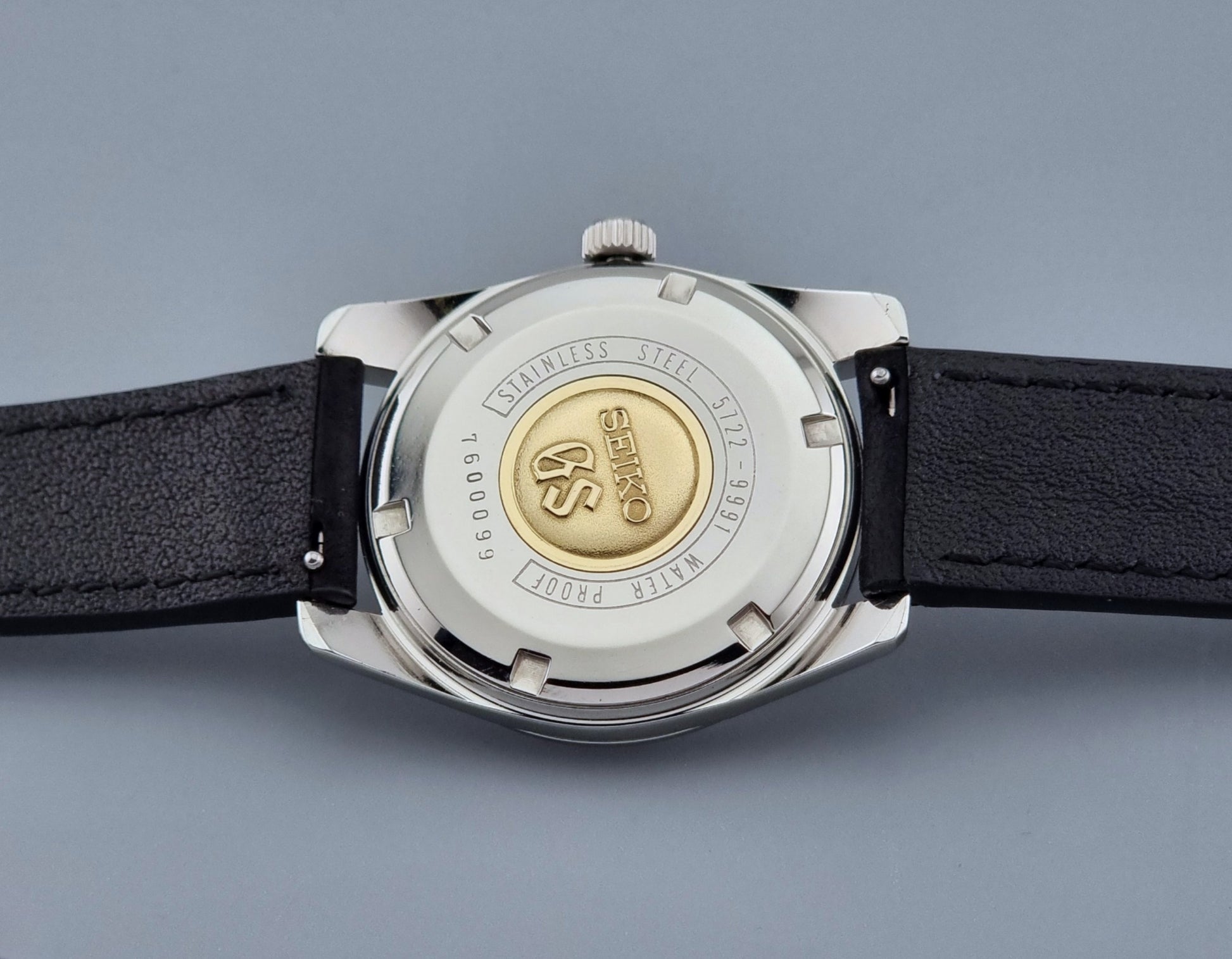 Vintage Grand Seiko Wristwatch 5722-9991 with black leather strap on a gray background case back with gold medallion showing the Seiko GS logo