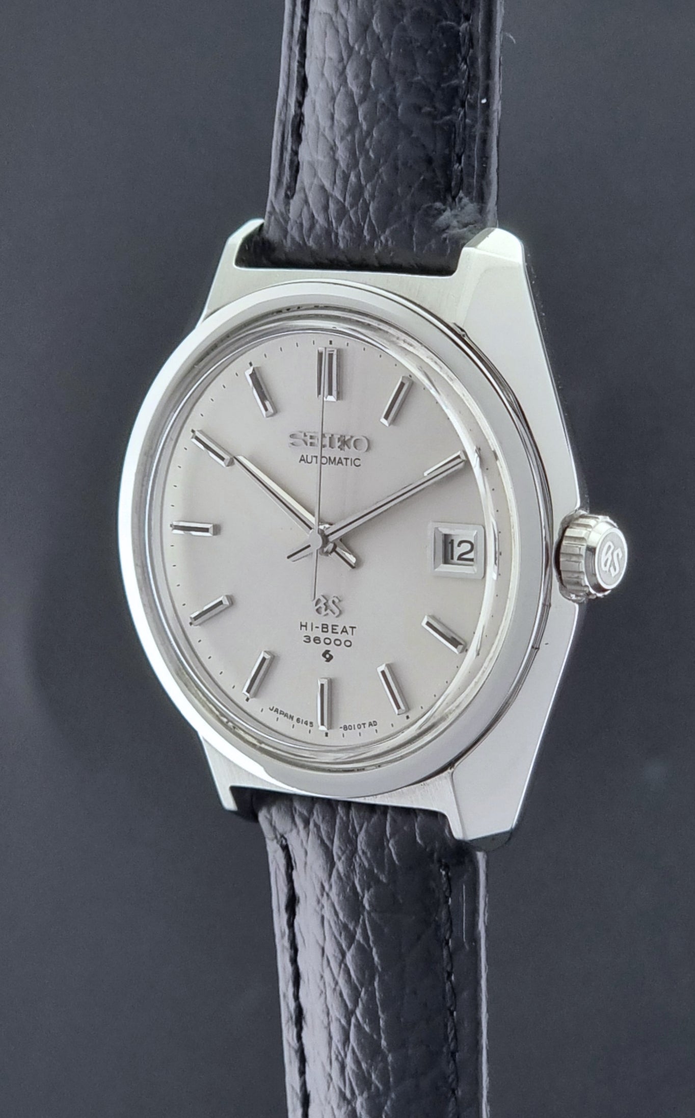 Grand Seiko Wristwatch 6145-8000 with black leather strap on a gray surface crown prolife view