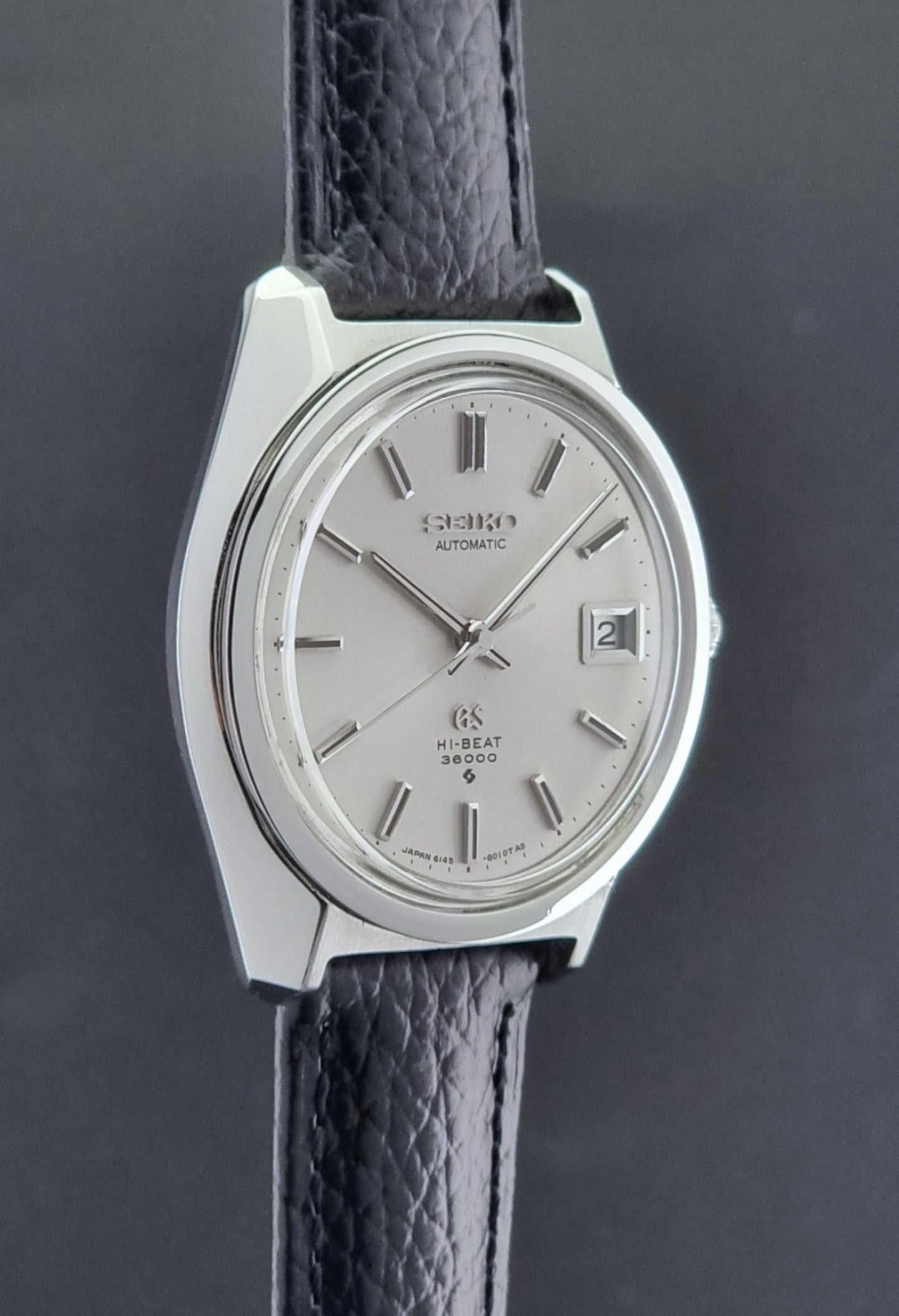 Grand Seiko Wristwatch 6145-8000 with black leather strap on a gray surface side profile view