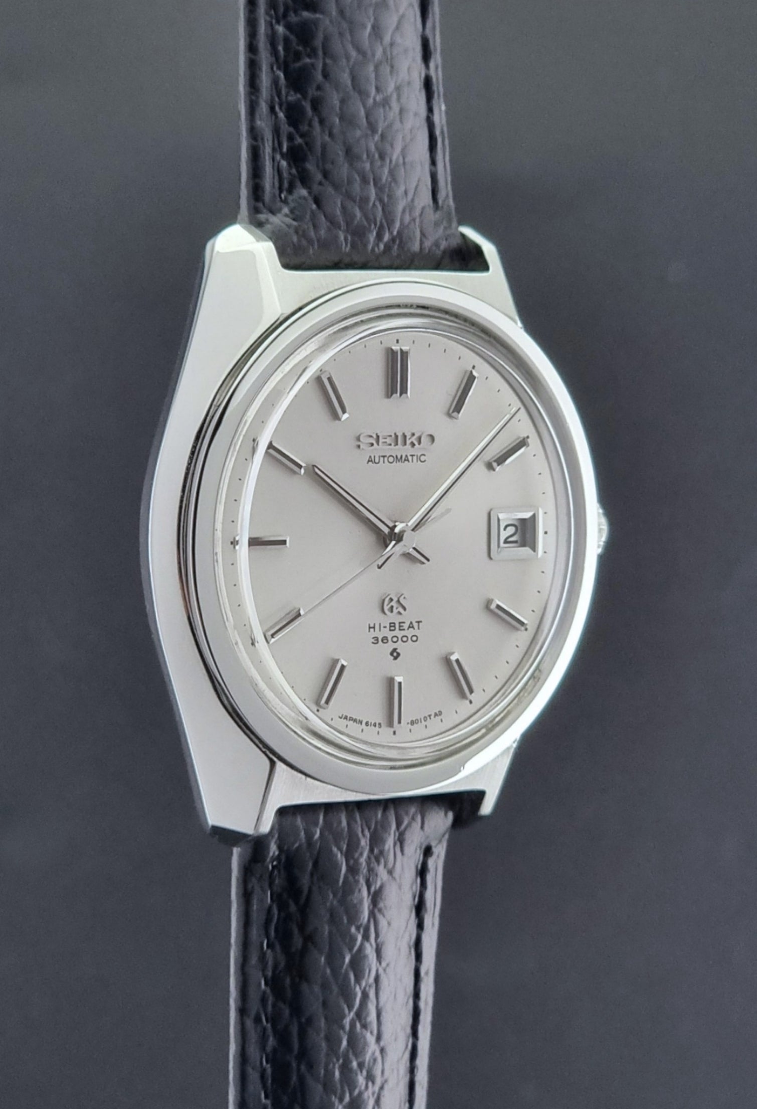 Grand Seiko Wristwatch 6145-8000 with black leather strap on a gray surface side profile view