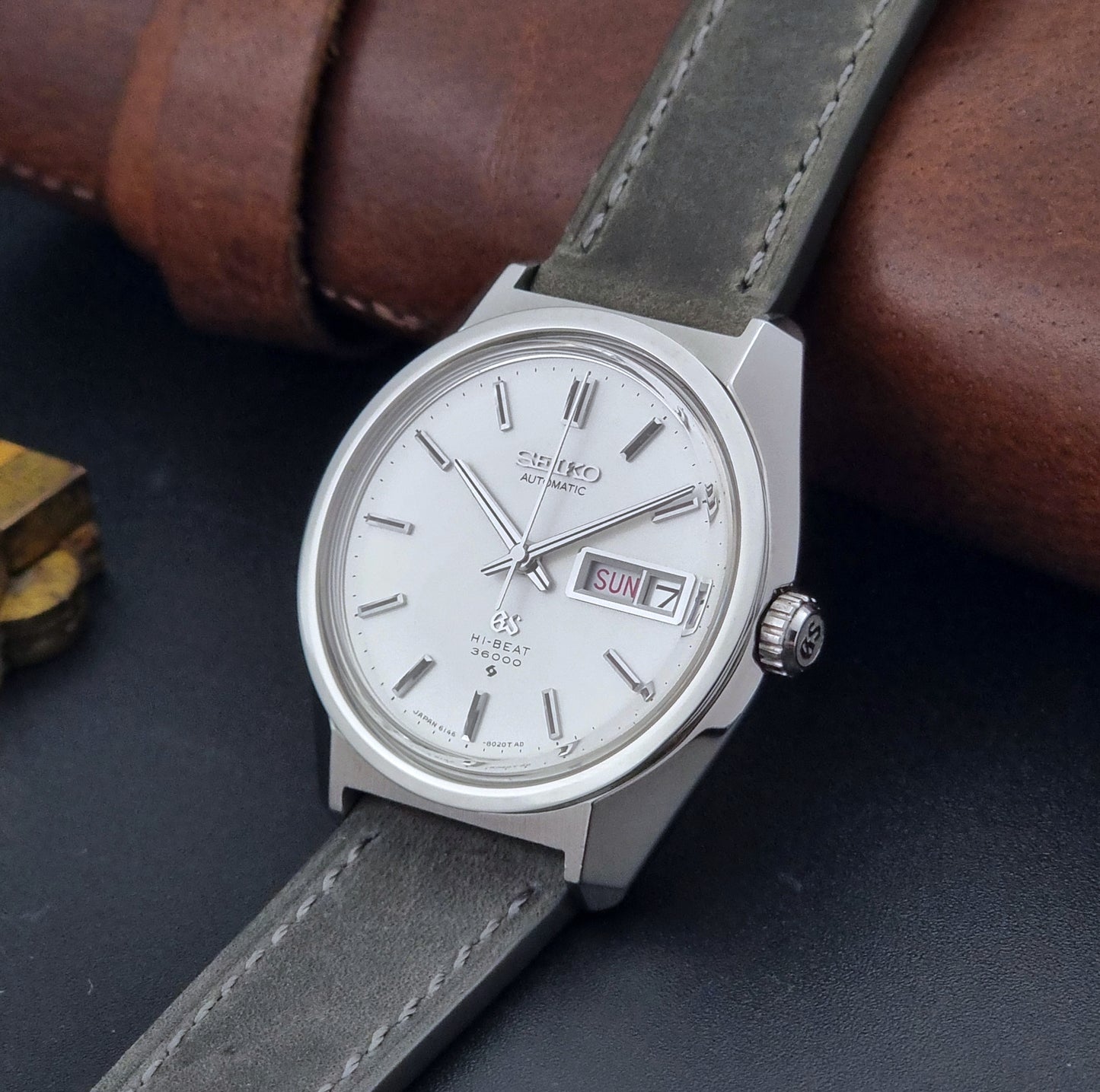 Vintage Grand Seiko Wristwatch 6146-8000 with gray strap and silver case on a dark grey background crown view