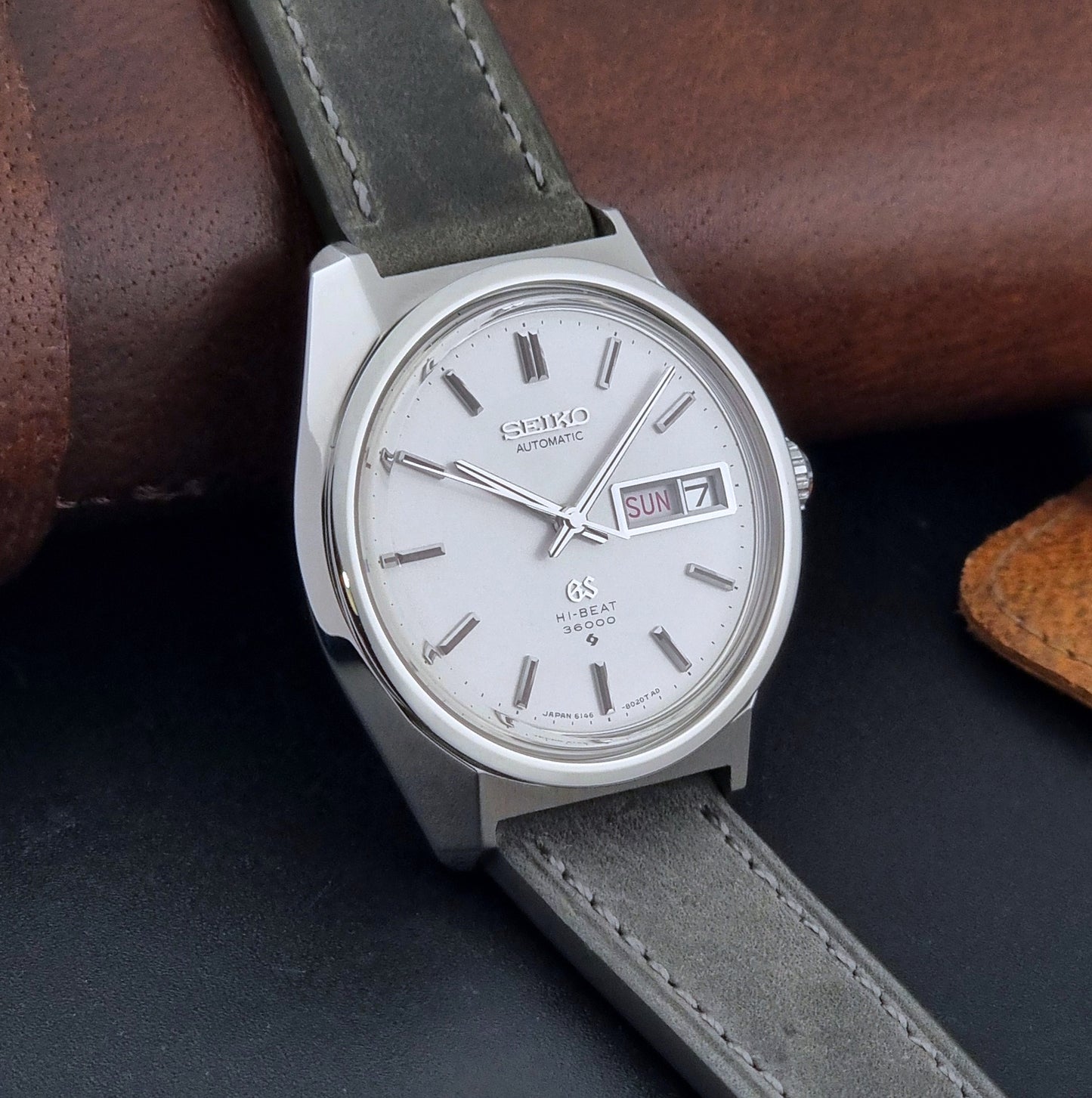 Vintage Grand Seiko Wristwatch 6146-8000 with gray strap and silver case on a dark grey background side view