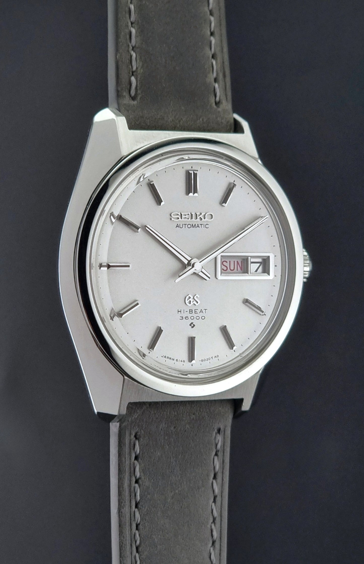 Vintage Grand Seiko Wristwatch 6146-8000 with gray strap and silver case on a dark grey background side profile view