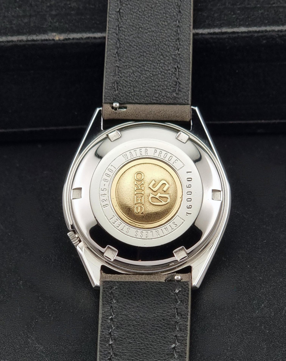 The back of a wristwatch with a silver stainless steel case and a dark gray leather strap, resting on a black textured surface. The case features a gold-colored central seal with the Seiko logo.