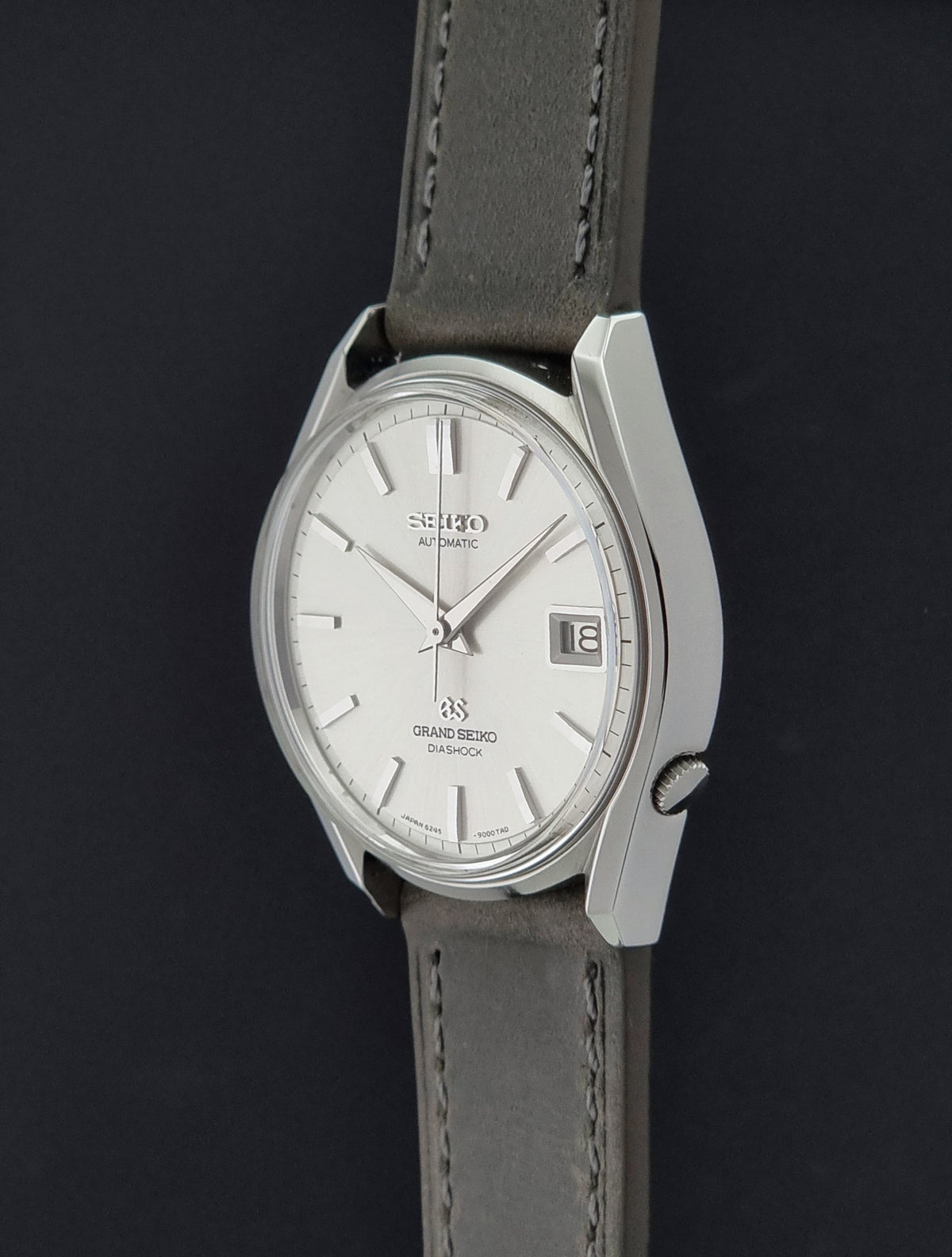 Vintage Grand Seiko 6245-9001 Watch featuring a silver-toned stainless steel case, a white dial with stick indices, and a dark gray leather strap, shown against a dark background.