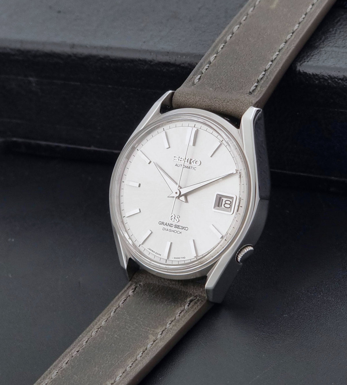 Vintage Grand Seiko 6245-9001 Watch with a silver-toned case, white dial, and a grey leather strap, resting on a dark textured surface.