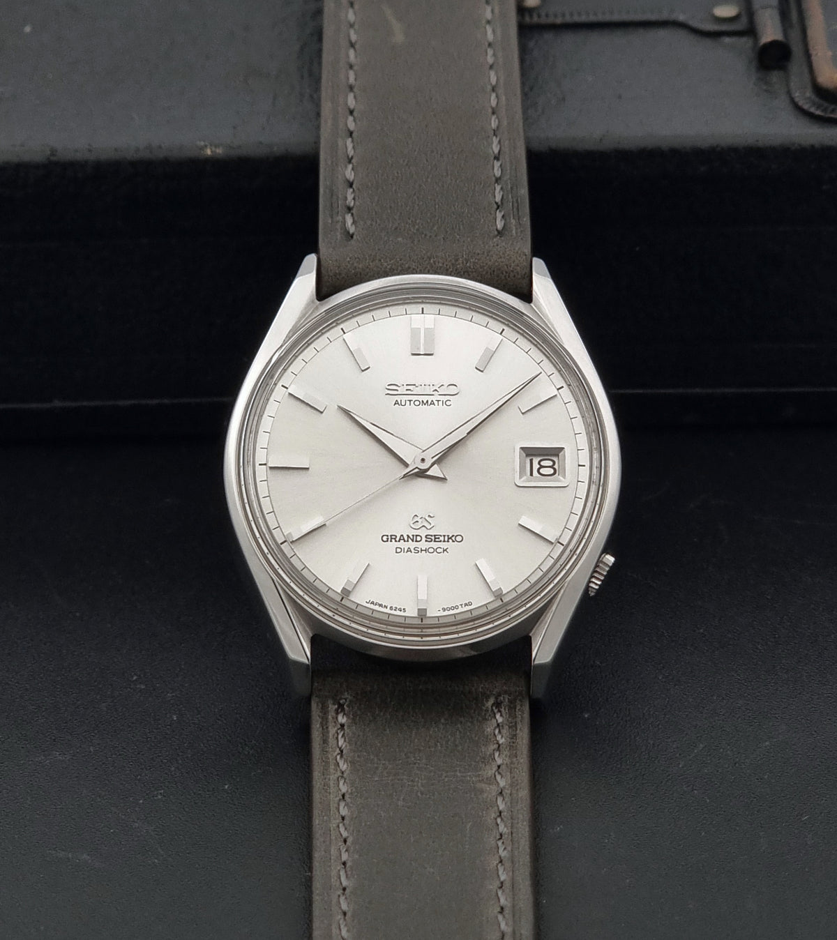 Vintage Grand Seiko 6245-9001 Watch with a silver dial, silver case, and a dark grey leather strap, displayed on a dark textured surface.