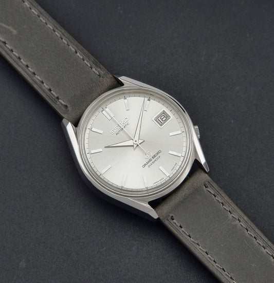Vintage Grand Seiko 6245-9001 Watch with a silver-toned oval case, white dial, date window, and dark gray leather strap, photographed on a dark textured background.