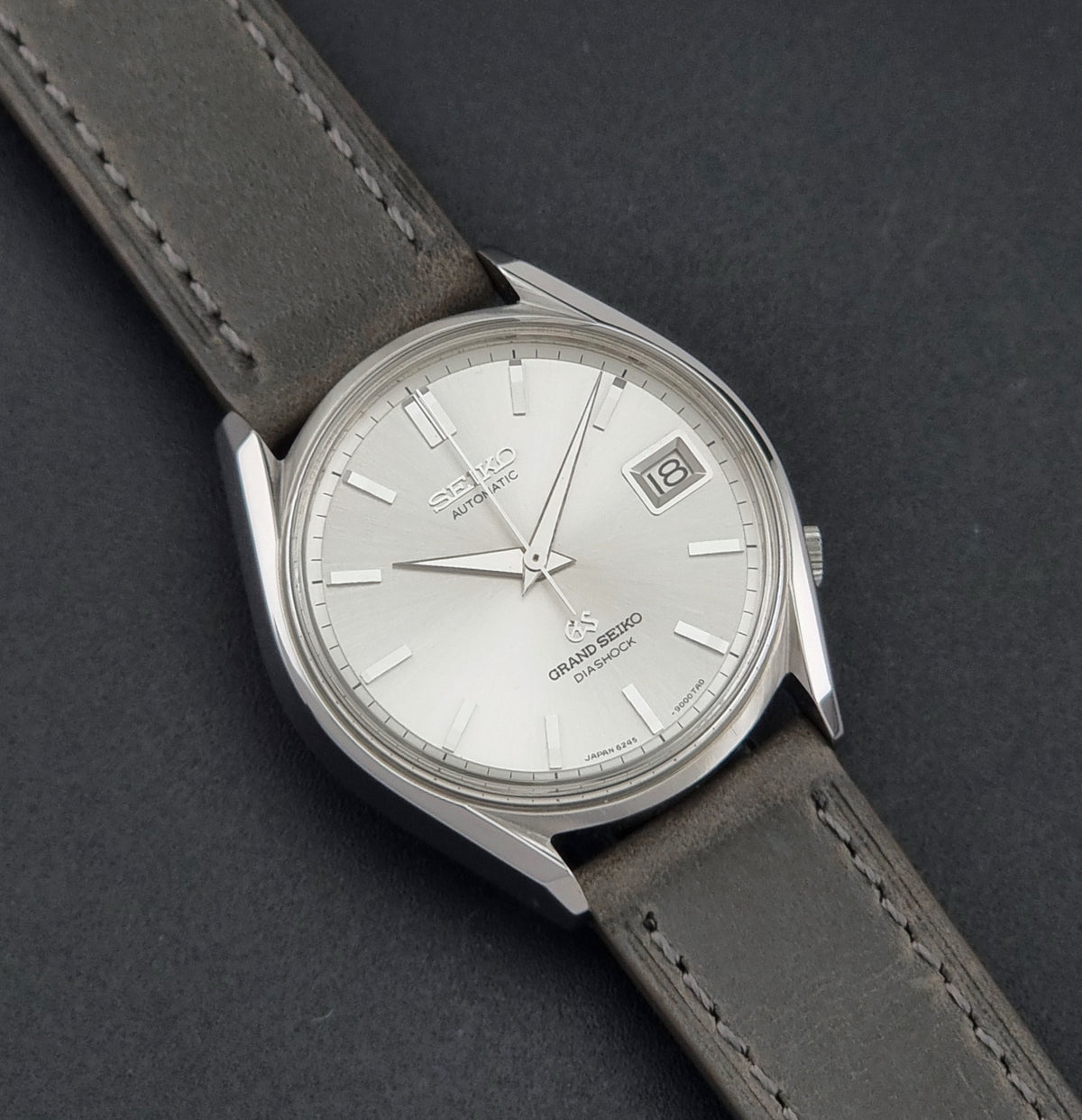 Vintage Grand Seiko 6245-9001 Watch with a silver dial, stainless steel case, and a dark gray leather strap, shown on a dark gray textured background.