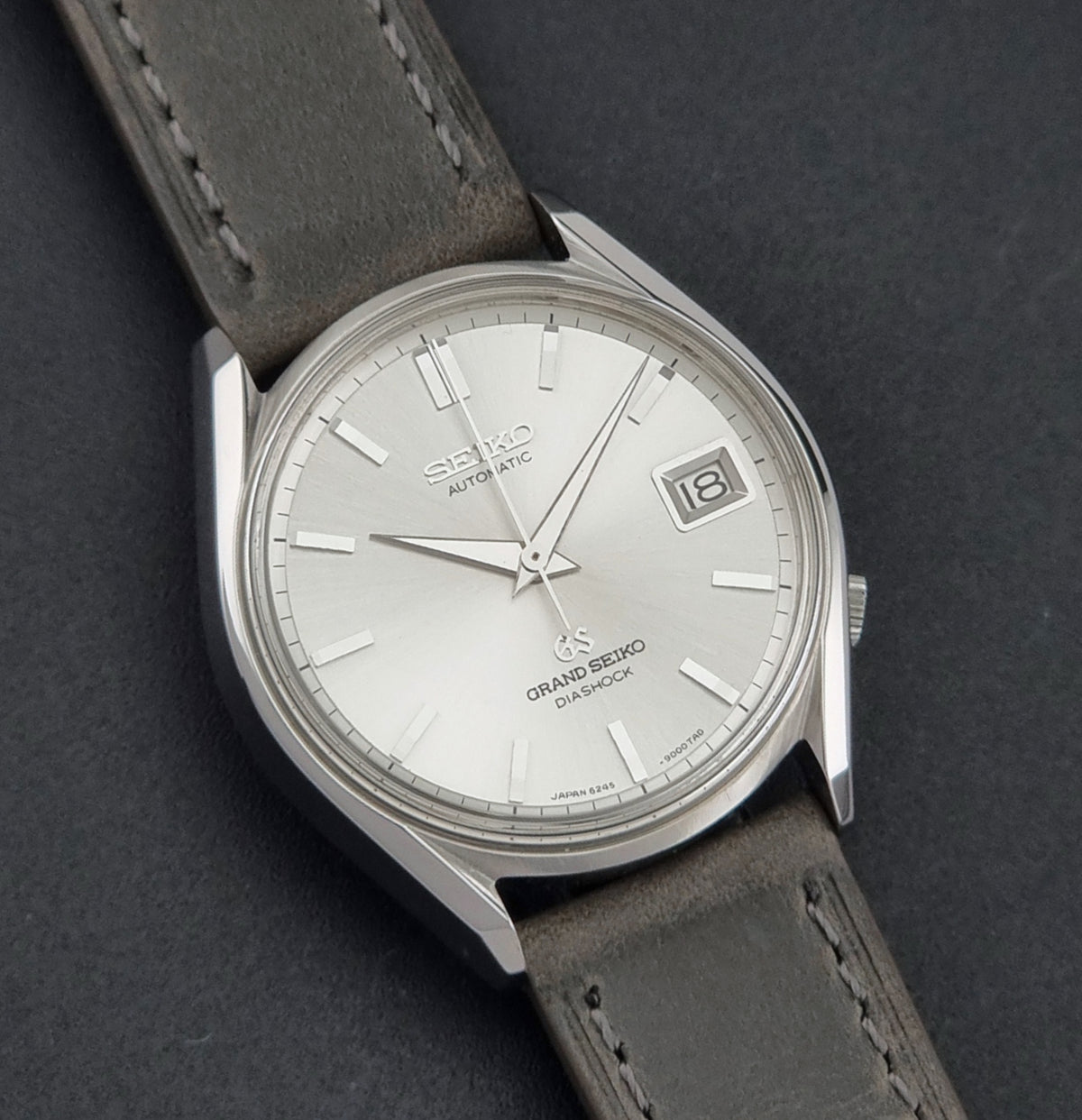 Vintage Grand Seiko 6245-9001 Watch with a silver dial, silver-tone case, and a dark gray leather strap, shown on a dark gray textured background.
