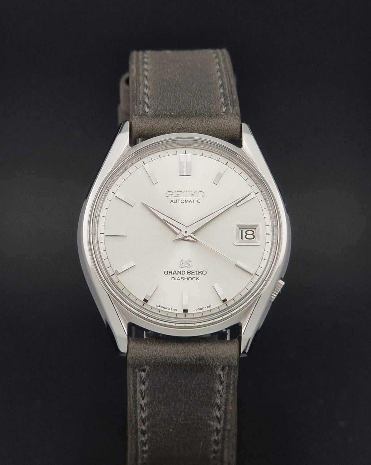 Vintage Grand Seiko 6245-9001 watch with a silver-toned stainless steel case, white dial, and a grey leather strap, shown against a dark background.