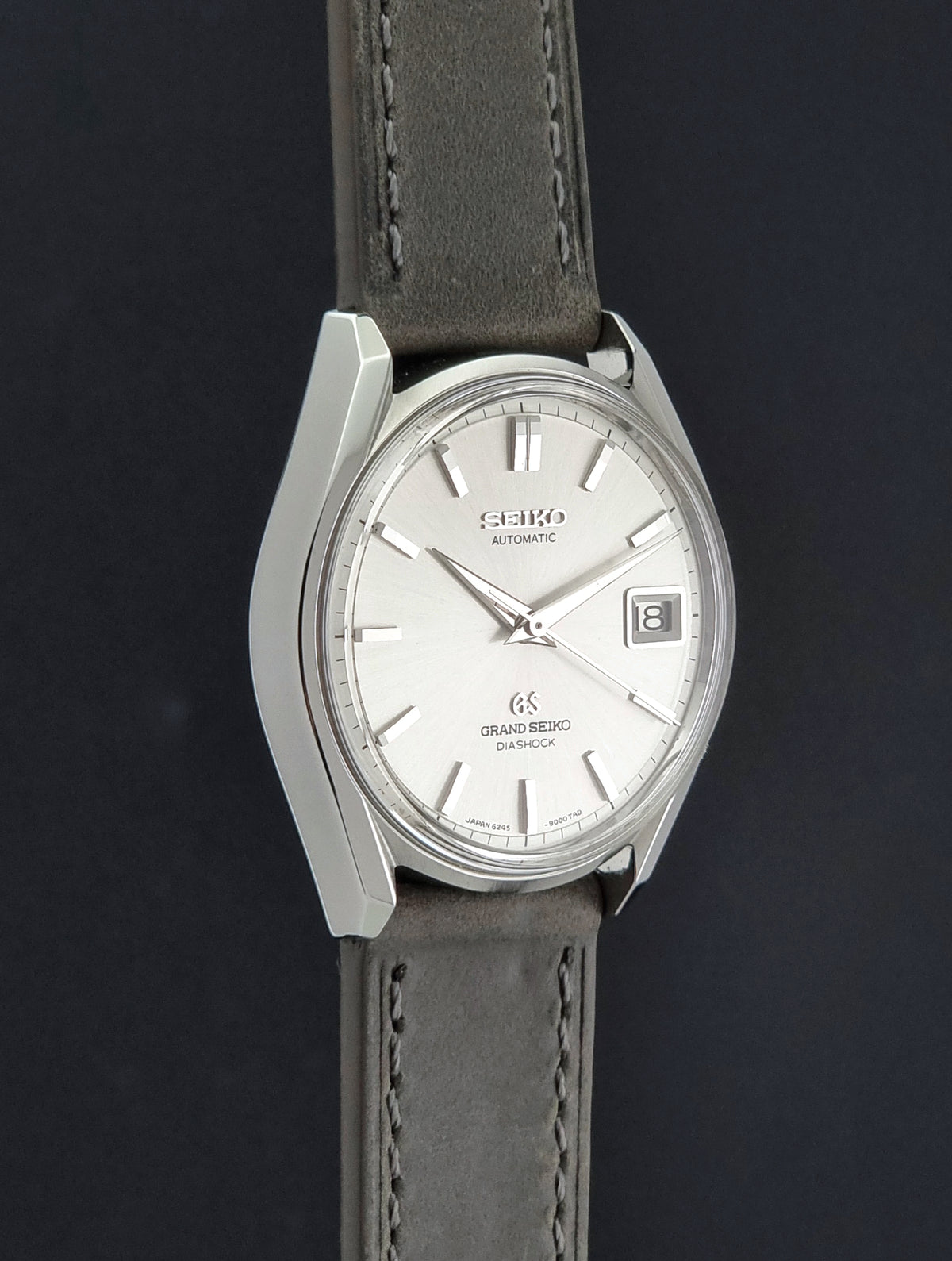 Vintage Grand Seiko 6245-9001 Watch with a silver-toned metal case, white dial, and a dark gray leather strap, shown against a dark background.