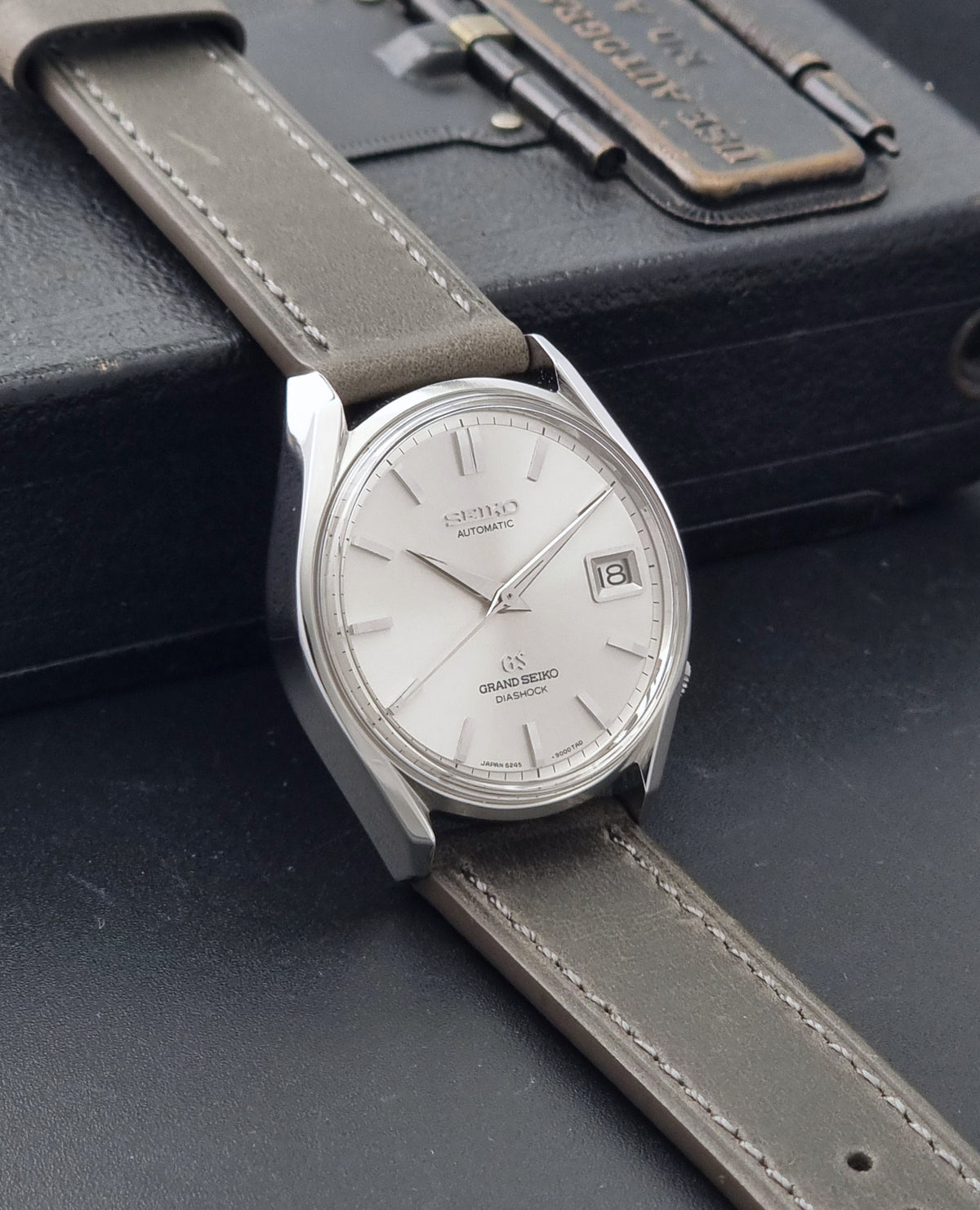 Vintage Grand Seiko 6245-9001 Watch with a silver-toned stainless steel case, white dial, and grey leather strap, resting on a dark textured surface,