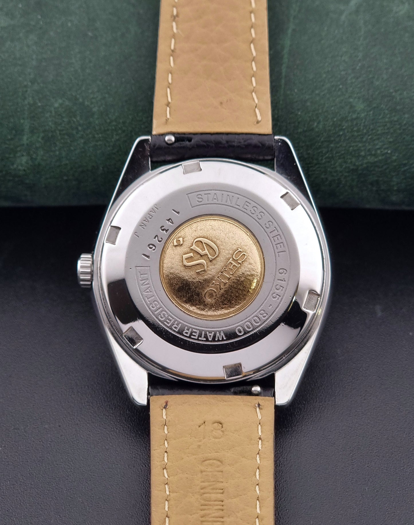 Vintage Grand Seiko Special 6155-8000 Silver wristwatch with black leather strap on a green and gray background case back view