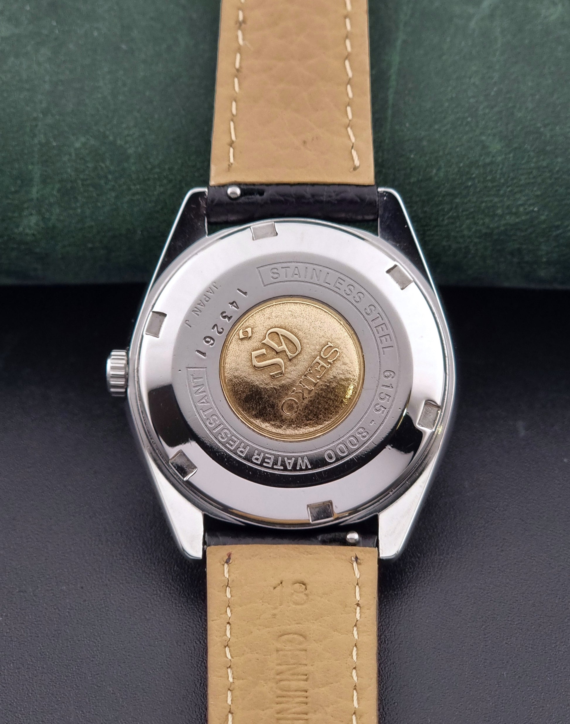 Vintage Grand Seiko Special 6155-8000 Silver wristwatch with black leather strap on a green and gray background case back view