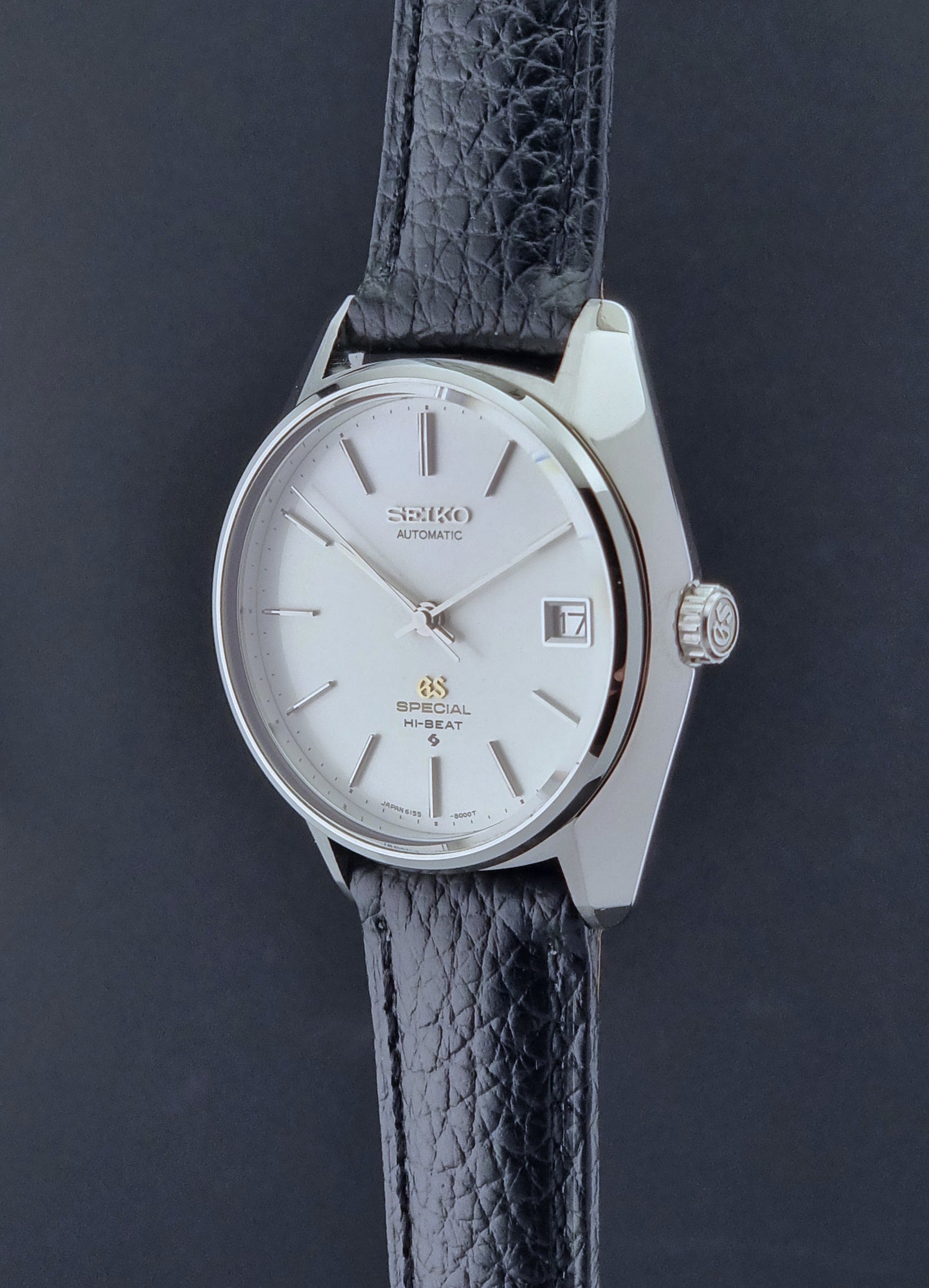 Vintage Grand Seiko Special 6155-8000 Silver wristwatch with black leather strap on gray background crown profile view