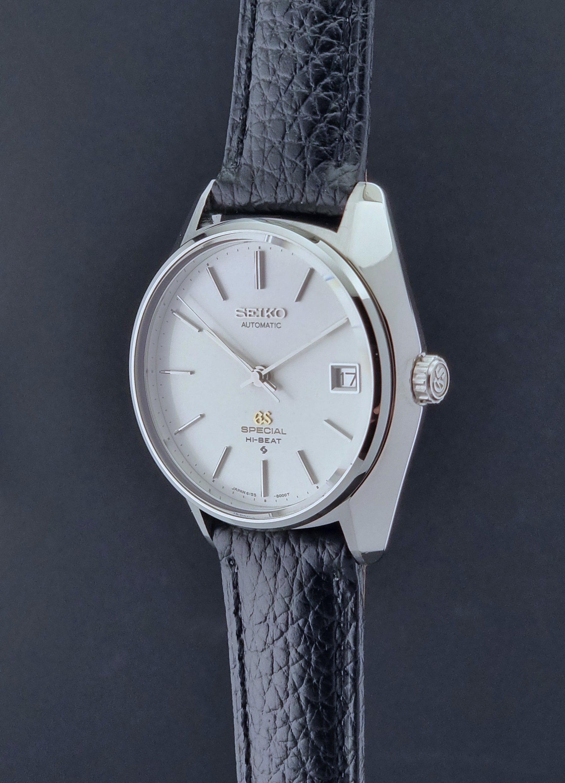 Vintage Grand Seiko Special 6155-8000 Silver wristwatch with black leather strap on gray background crown profile view